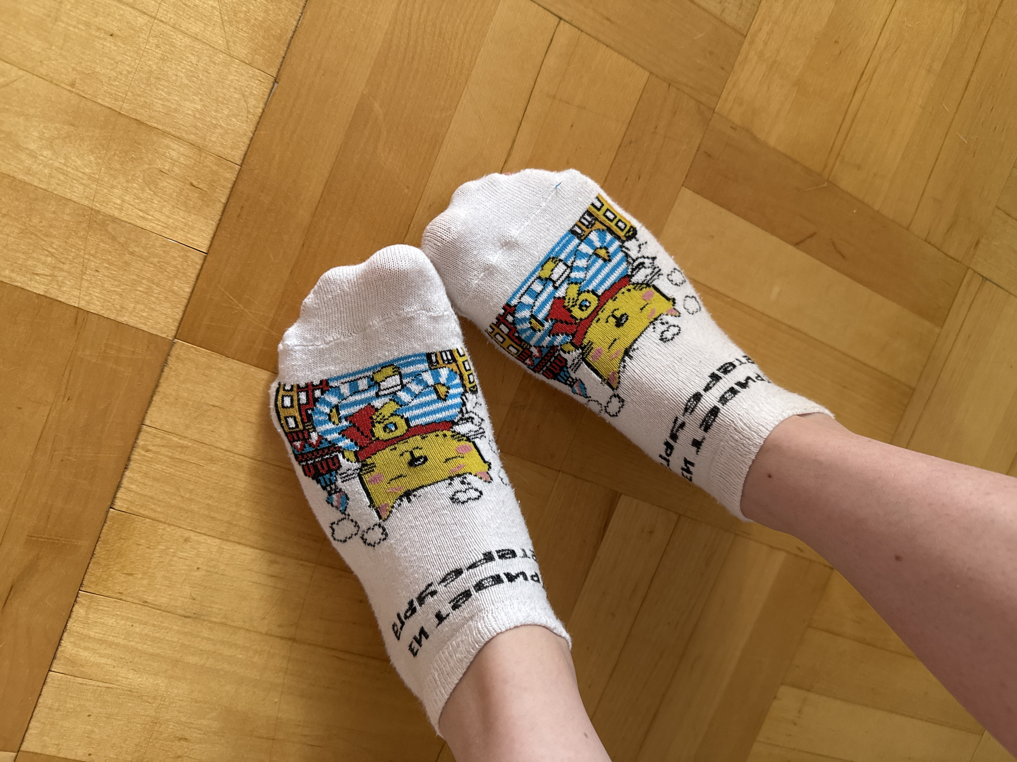 The image displays a top-down view of a person's lower legs and feet, adorned with white ankle socks, resting on a warm, light brown wooden floor. The floorboards are arranged in a geometric pattern, possibly herringbone or parquet. The person has fair skin.
Both socks feature a vibrant cartoon design. Each sock showcases a yellow, animal-like character, reminiscent of Pikachu or a similar creature, dressed in red and blue striped pajamas and holding what appears to be a pillow or plush toy. The background of the cartoon design has matching blue and white stripes. Scattered around the main character are small white outlines, possibly depicting clouds or bubbles.
Cyrillic text is visible on the ankle cuffs of both socks, printed upside down from the viewer's perspective. The text reads "ПРИВЕТ ИЗ САНКТ-ПЕТЕРБУРГА," which translates to "Greetings from Saint Petersburg."
The scene depicts an indoor setting, likely with the person relaxing or standing. The lighting suggests it is daytime. The explicit location is unknown, though the socks themselves offer a geographical cue.