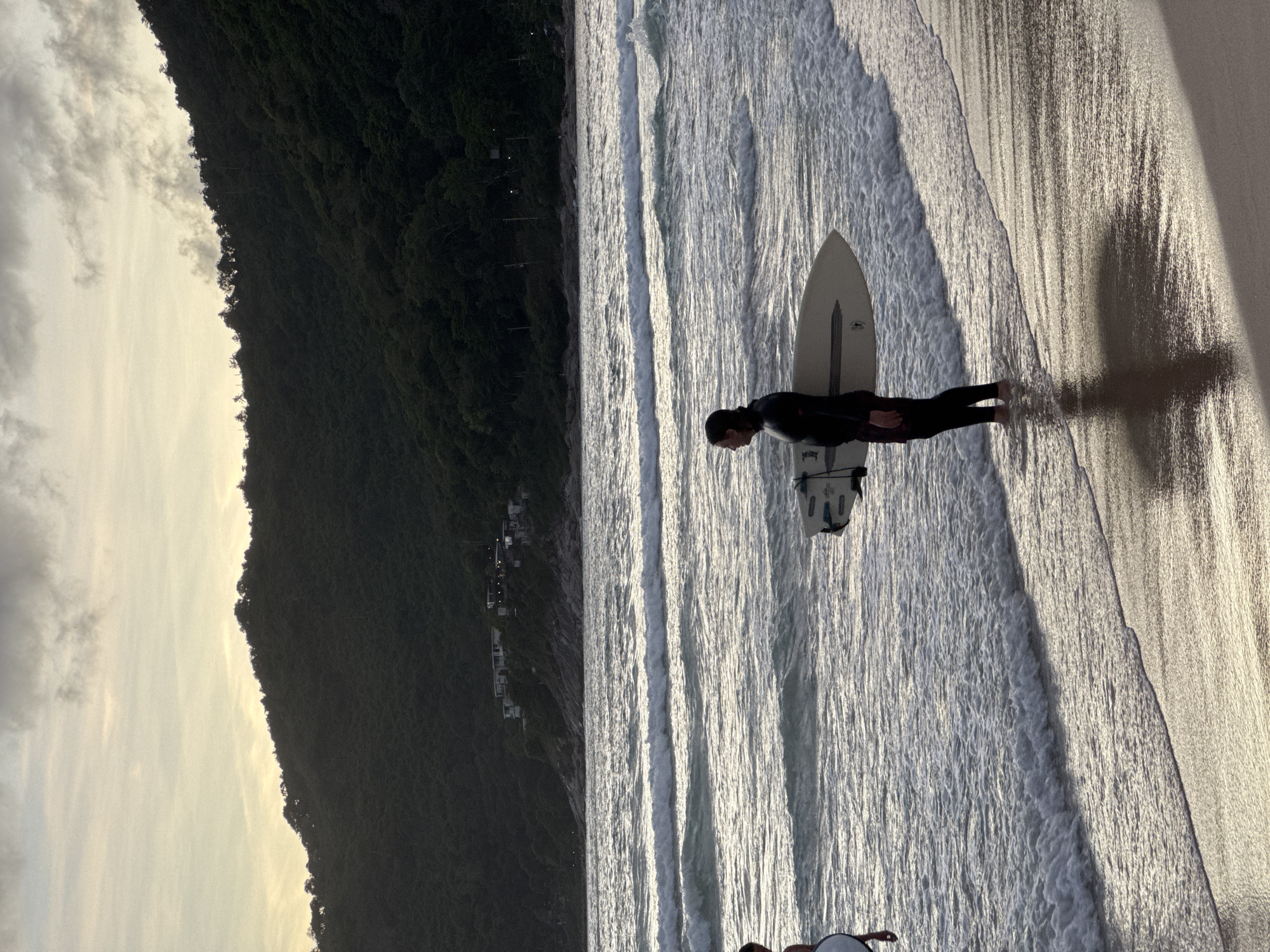 A lone surfer stands in shallow water on a sandy beach, holding a surfboard under their right arm. The surfer is wearing a black wetsuit with red patterned shorts underneath. The water is calm with small waves breaking on the shore. In the background, a dense, tree-covered hillside rises, with a few buildings visible on the slope. The sky above is overcast with a pale yellow hue, suggesting either sunrise or sunset. The scene is set in Rawai, Thailand.