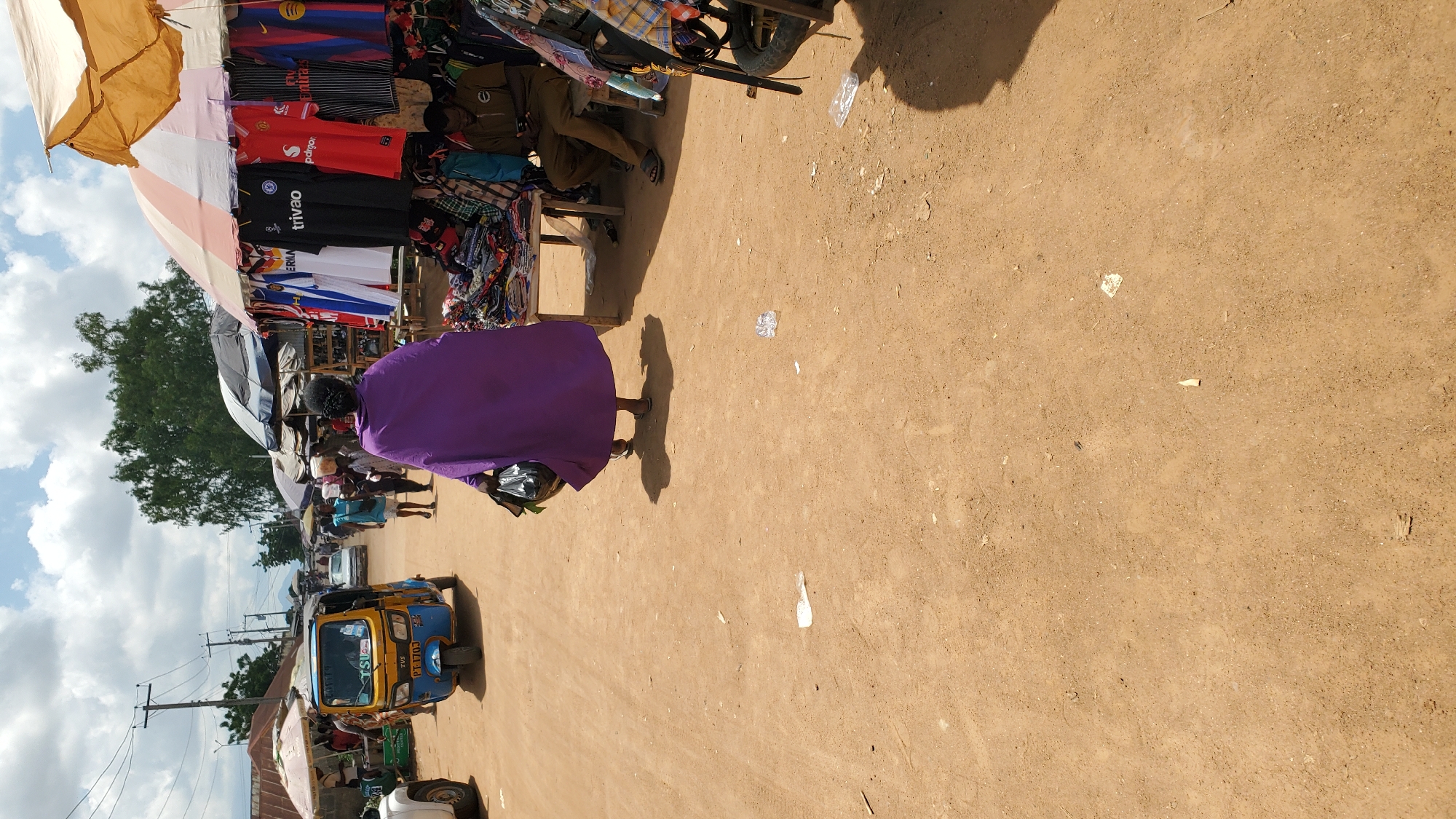 The image depicts a bustling outdoor market scene in Jalingo, Nigeria. In the foreground, a person walks away from the camera, draped in a vibrant purple garment and carrying a black plastic bag. The ground is dusty and uneven, with scattered debris. To the right, a market stall is set up under a large umbrella, displaying various colorful t-shirts. A man sits on a low bench to the right of the stall, looking down at his phone. In the background, a blue and yellow auto-rickshaw (Keke Napep) is parked on a dirt road, with other market-goers and stalls visible further down the street. The sky is partly cloudy, indicating a bright, sunny day. The overall impression is one of vibrant commerce and daily life in a Nigerian city.