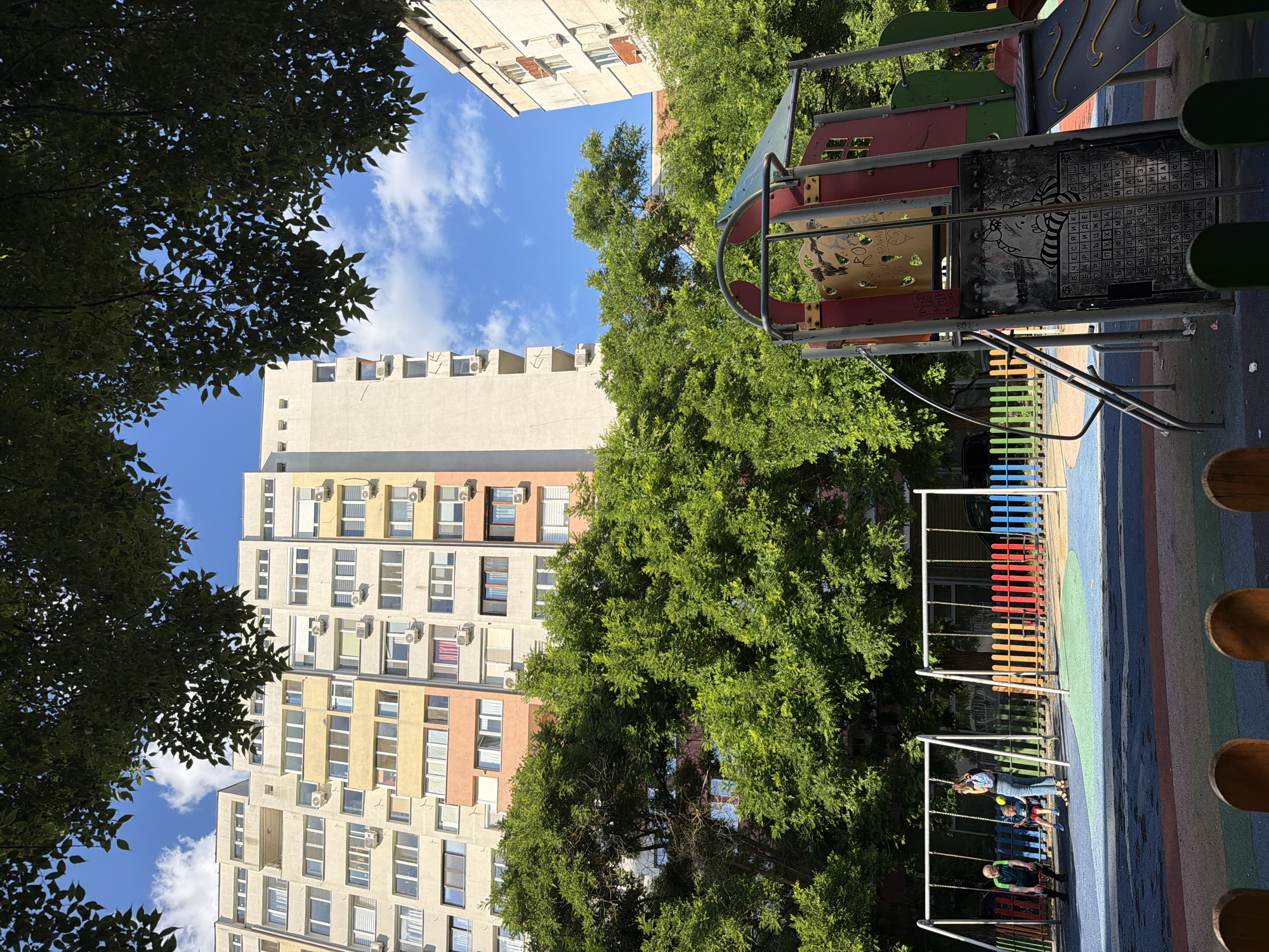 This image captures a playground area in the city of Burgas, Bulgaria, situated at the base of multi-story residential buildings. The foreground features a colorful playground with swings and a climbing structure, partially obscured by wooden fencing. In the mid-ground, a woman is pushing a child on a swing, while another person sits on a separate swing set. Lush green trees form a dense canopy, separating the playground from the imposing apartment blocks visible in the background. The buildings are a mix of light-colored facades with numerous windows and air conditioning units, indicative of a typical urban residential area. The sky is blue with scattered white clouds. The scene suggests a sunny day in a densely populated urban environment, with the playground offering a recreational space amidst the concrete structures.
