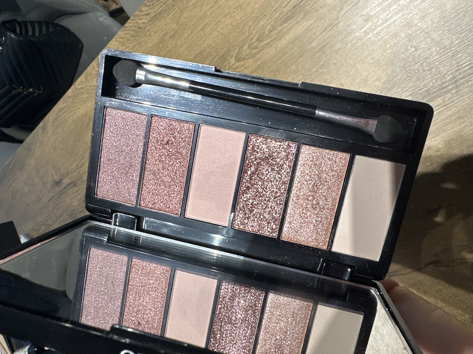 A close-up photograph shows an open eyeshadow palette on a wooden surface. The palette has seven rectangular pans of eyeshadow in varying shades of pink and beige, some with a glitter finish. A dual-ended eyeshadow applicator sits on top of the pans. The palette is open, revealing a mirrored surface showing a reflection of the eyeshadows. The wooden surface appears to be part of a table. The palette is held open with a hand. The location in the city of Plovdiv, Bulgaria, is not discernible from the image.