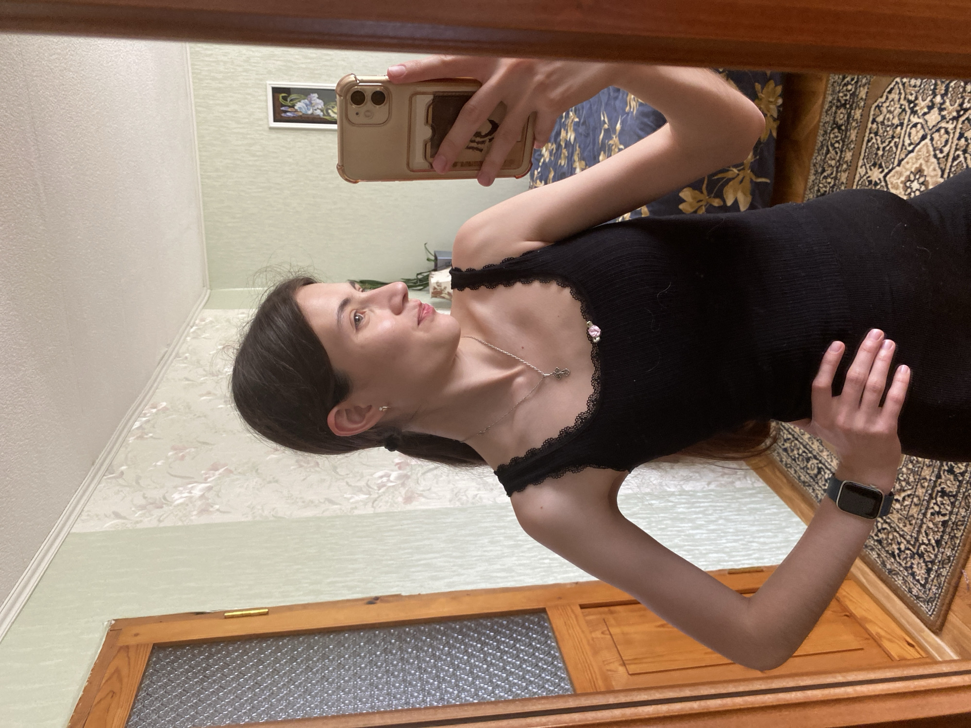 A young woman with dark hair pulled into a ponytail, wearing a black ribbed tank top with lace trim, takes a mirror selfie in what appears to be a residential interior. She is positioned slightly to the left of the frame, looking towards her right. She is wearing a silver necklace with a cross pendant and a watch on her left wrist. Her right hand is placed on her hip, and her left arm is slightly bent. The mirror reflects the room behind her, which includes a wooden door with frosted glass on the left, patterned wallpaper with a pale green base and pink floral designs, and a bed with a dark blue comforter adorned with yellow floral motifs. A framed picture of blue flowers hangs on the wall. The floor is covered by a patterned rug. The lighting suggests an indoor setting during the daytime.