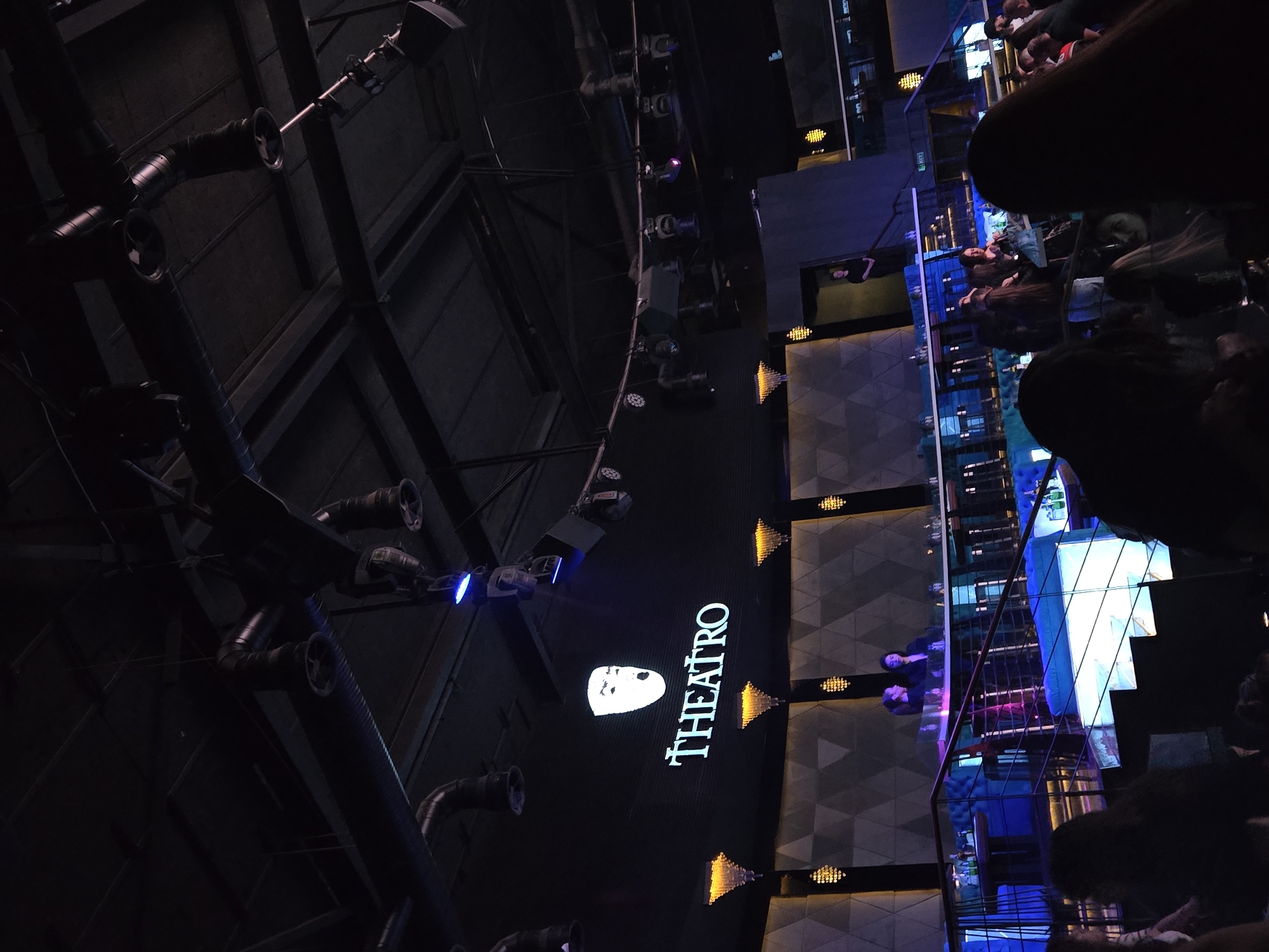 The photograph depicts the interior of a venue called "THEATRO" in an unknown city, likely a nightclub or theater. The image is taken from a low angle, looking upwards. The architecture features industrial-style piping and a high ceiling. On the right, there are multiple levels of the venue with people sitting at tables. The lighting is a mix of blue and yellow, creating a vibrant atmosphere.