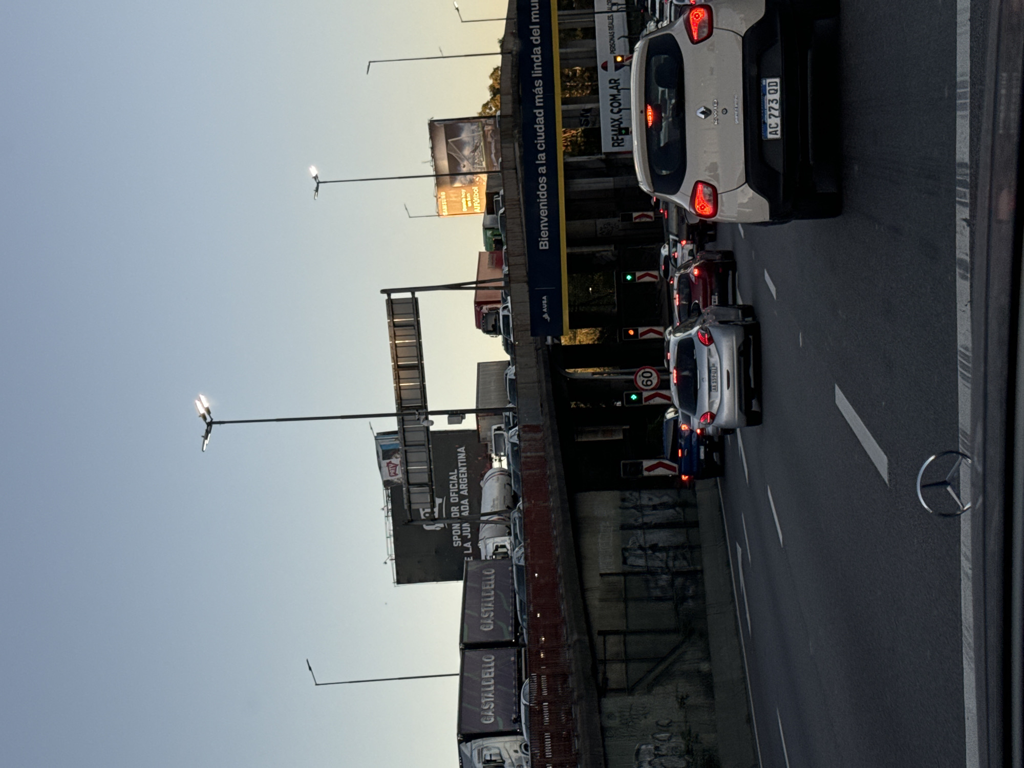 Here's a detailed description of the image:
**Setting:** A highway in Buenos Aires, Argentina. The photograph is taken from inside a vehicle, likely a bus or car, in the middle of heavy traffic. The sky is a clear blue, indicating a day with good weather. The time of day seems to be either early morning or late afternoon, based on the angle of the sunlight.
**Objects:**
* Many cars are congested on the highway, ranging in color from white, silver, blue, and red.
* A white Renault Kwid with the license plate "AC 273 QD" is directly in front.
* Billboards line the side of the road, with ads for companies like "Castaldello," "Al Futbol," and "Samsung."
* There is a large overhead sign that welcomes visitors to the city, stating "Bienvenidos a la ciudad más linda del mu".
* Street lamps are spaced out along the highway.
* There is a speed limit sign with the number 60.
* A truck with the "GASTALDELLO" logo is also visible.
* A logo of a Mercedes-Benz is located on the right side of the image.
**Subjects/Actions:**
* The primary subject is the traffic congestion on the highway.
* Cars are either stopped or moving slowly.
* The photograph appears to be taken by a passenger inside one of the vehicles.
**Environment:**
* The surrounding environment is urban, indicated by the infrastructure and billboards.
* The road appears to be well-maintained.
* A concrete wall runs along the side of the road, possibly as a safety barrier.