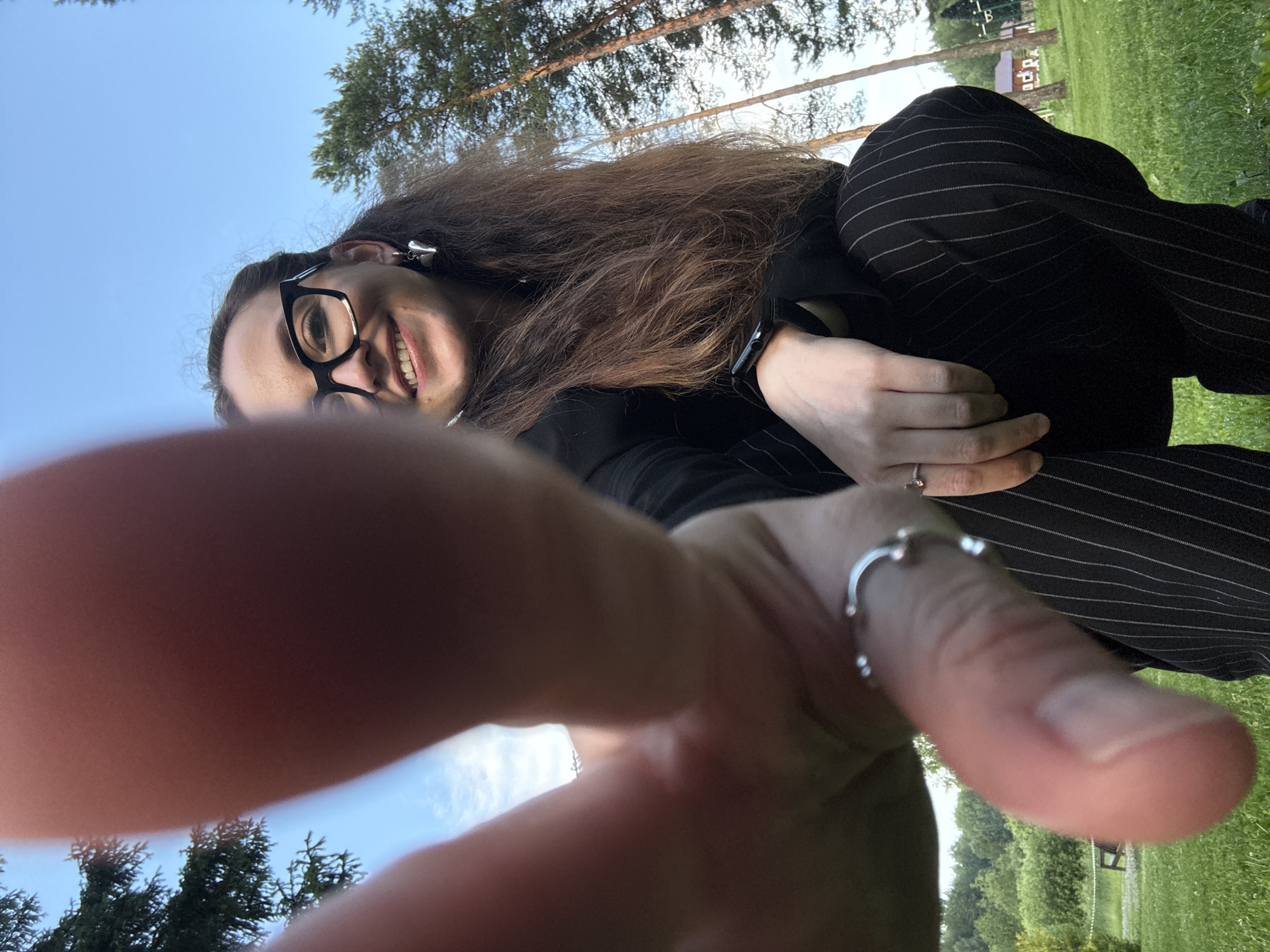 The image shows a close-up, low-angle shot of a person with long brown hair, wearing glasses and a black outfit with pinstripes. The person is smiling at the camera. Their left hand is in the foreground, partially obscuring the camera's view, with rings on their fingers. The background appears to be outdoors, with trees and a grassy area visible. The sky is blue, suggesting it is daytime, likely sunny. The overall impression is of a selfie taken in a park or garden setting. The location is identified as Votkinsk, Russia.