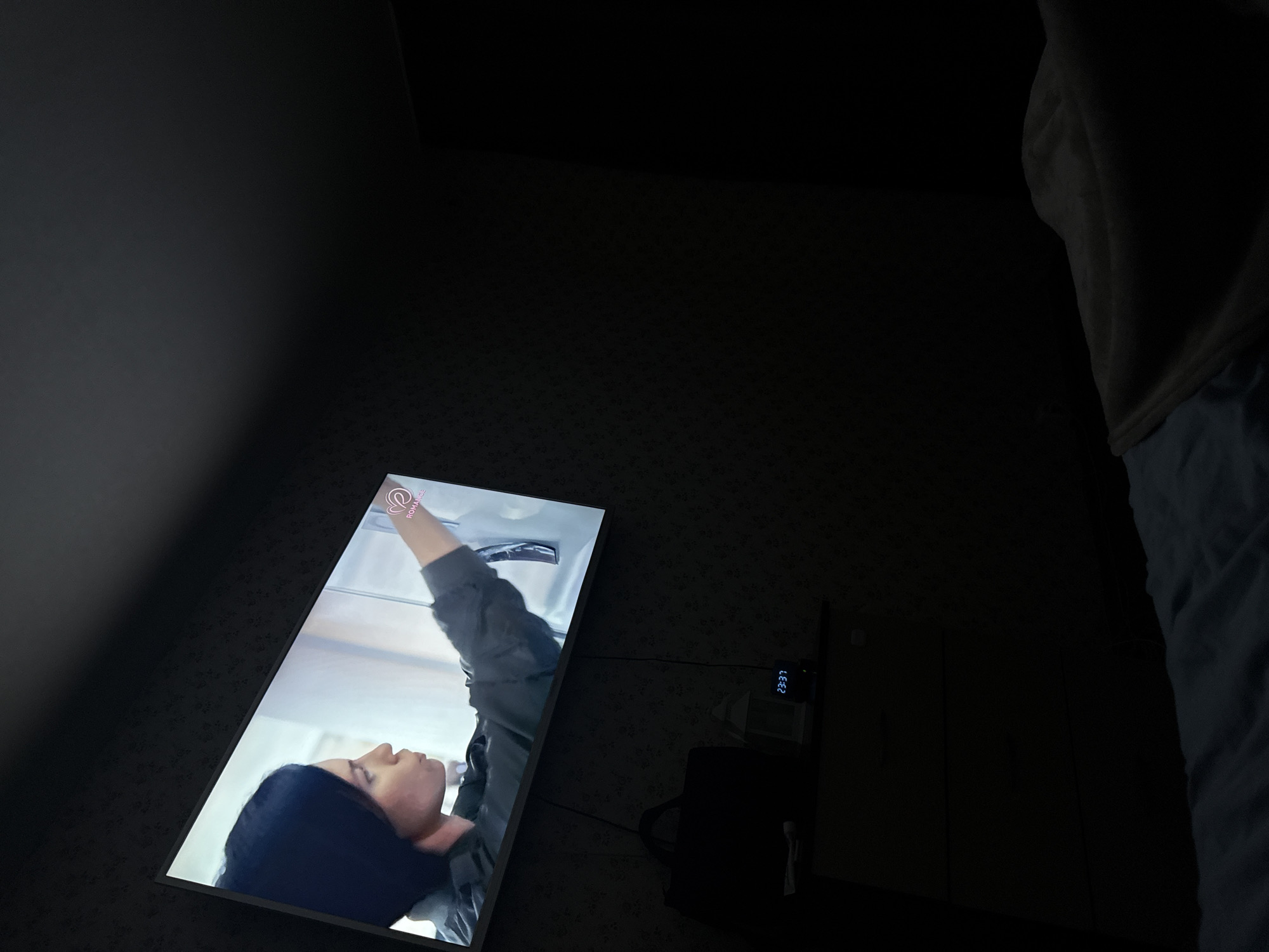 A low-lit interior room scene features a television screen displaying a woman in profile, reaching up, possibly in a scene from a show titled "Romance." To the right of the screen is a nightstand with a digital clock displaying "23:37," along with a dark, obscured object that could be a bag. The edge of a bed with white bedding is visible on the right side of the frame. The room itself appears to have patterned wallpaper. Location information is not discernible.
