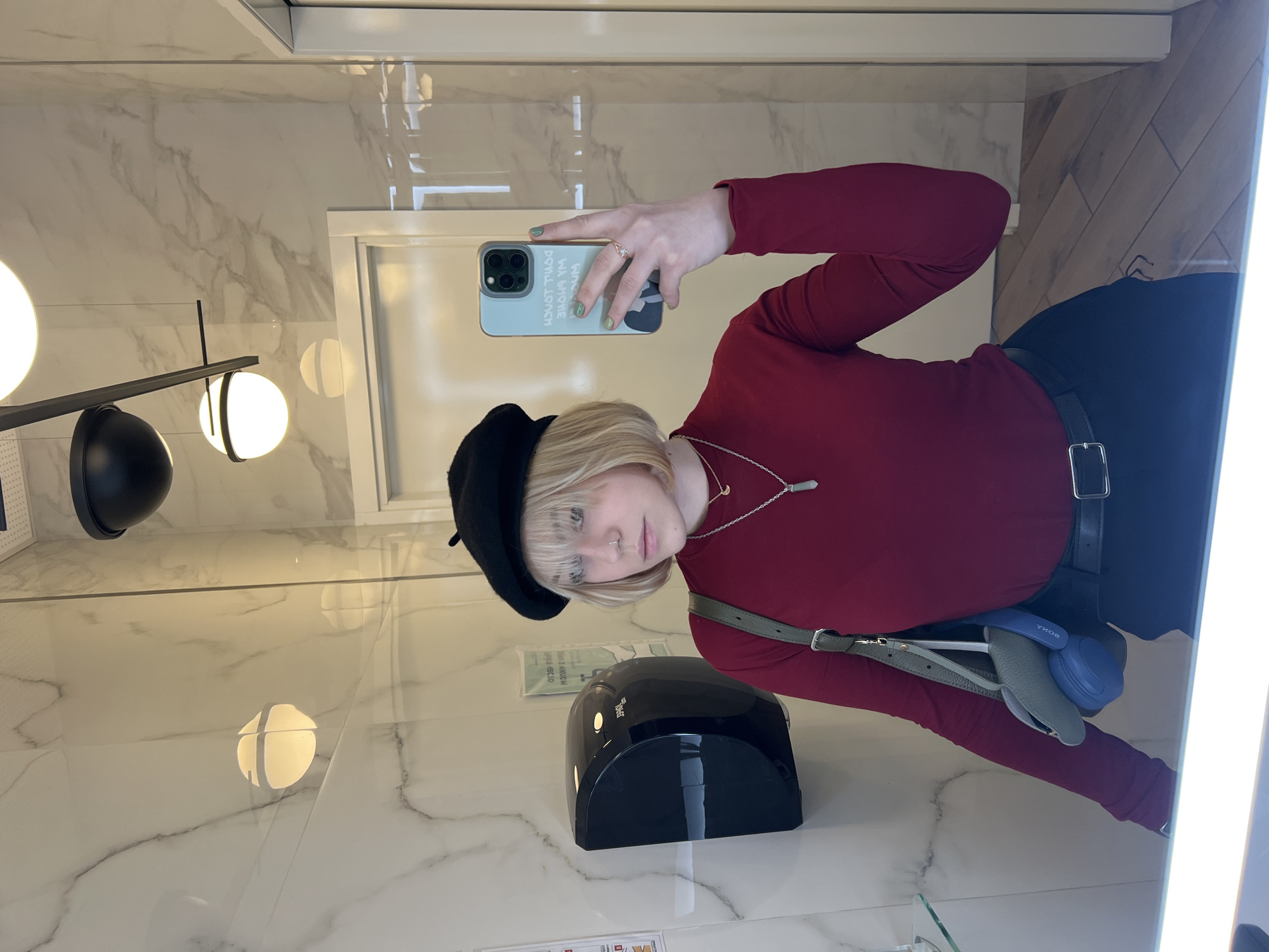 A young person, likely in a modern restroom in Odesa, Ukraine, captures a mirror selfie. They have short, light blonde hair with bangs, a nose piercing, and a neutral expression looking directly into the mirror. They are wearing a black beret, a long-sleeved dark red top, a black belt, and a silver necklace with a crystal pendant. Their right hand, adorned with light green nail polish and a ring, holds a light blue smartphone displaying reversed text "DON'T TOUCH MY PHONE" and "MY AUDIO". A grey-green shoulder bag is visible, with blue Sony over-ear headphones (brand "SONY" visible reversed) tucked into its strap.
The setting features glossy white wall tiles with grey marble-like veining. Modern lighting fixtures with multiple spherical white lamps and black accents are reflected throughout the mirror. A black hand dryer, partially revealing the reversed brand "DYSON," is mounted on the wall to the person's left. The floor on the right side of the image consists of light brown wooden-patterned tiles. Bright vertical strip lighting is reflected along the right edge of the mirror. The scene is well-lit, suggesting an indoor commercial or public space.