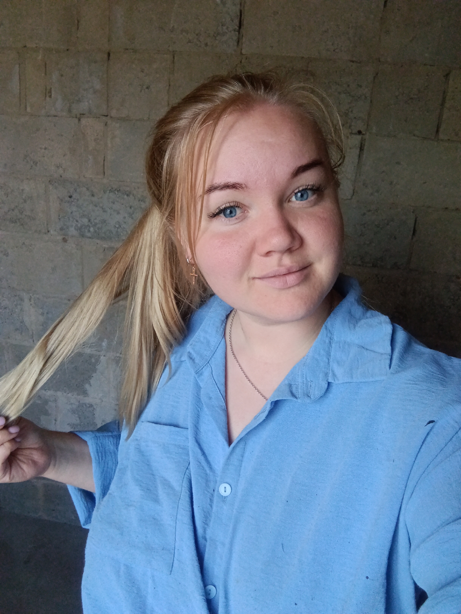 A young adult female is depicted in a chest-up portrait, facing forward and looking directly at the viewer. She has blonde hair, partially tied back in a ponytail, with strands framing her face, and blue eyes. Her complexion is fair with some visible redness on her cheeks and forehead. She is wearing a light blue, collared, short-sleeved shirt made of a textured fabric, with light blue buttons down the front and a visible left chest pocket. Small dark specks are present on the shirt's fabric, and a small tear or hole is visible on the upper right shoulder area of the garment. A thin, silver-colored chain necklace is visible around her neck, and a small, gold-colored cross earring is present in her right ear. Her right hand is positioned in the lower-left quadrant of the image, holding a section of her ponytail. The background consists of an unpainted, coarse-textured concrete or cinder block wall, exhibiting visible mortar lines and variations in gray tone, consistent with an unfinished or utilitarian structure. The scene is located in Фоки, Russia.