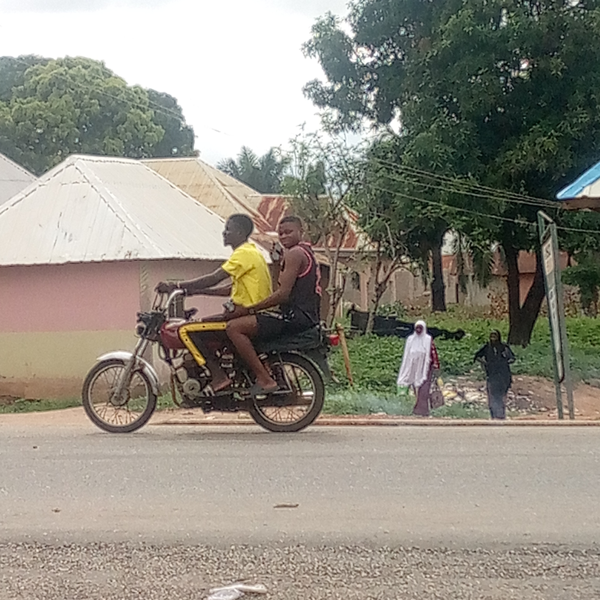 Two young men are riding a red motorcycle with a yellow stripe down a paved road. The driver is wearing a yellow shirt, and the passenger is wearing a black tank top. Two women are walking on the side of the road in the background. One is wearing a white hijab and the other a black top. Buildings and trees are visible in the background. The scene appears to be in Mararaba, Nigeria.