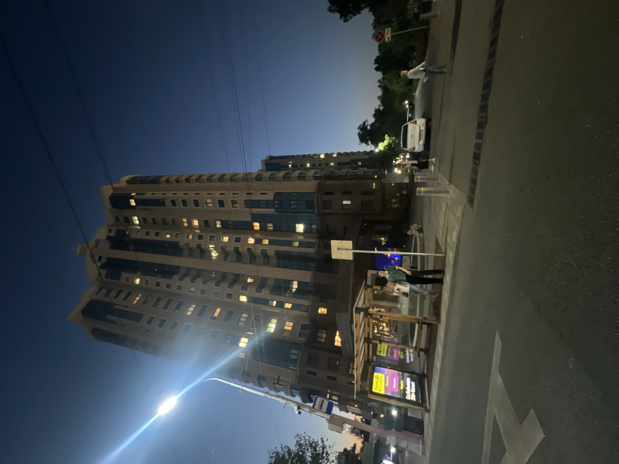 The image shows a street scene in Almaty, Kazakhstan, at dusk. A tall, modern apartment building with numerous illuminated windows dominates the background. In the foreground, a bus stop shelter with advertisements is visible, and several people are standing nearby. A street light illuminates the scene. A white car is parked on the street, and a traffic sign is visible on the right side of the image. The sky is a deep blue, indicating the transition from day to night.