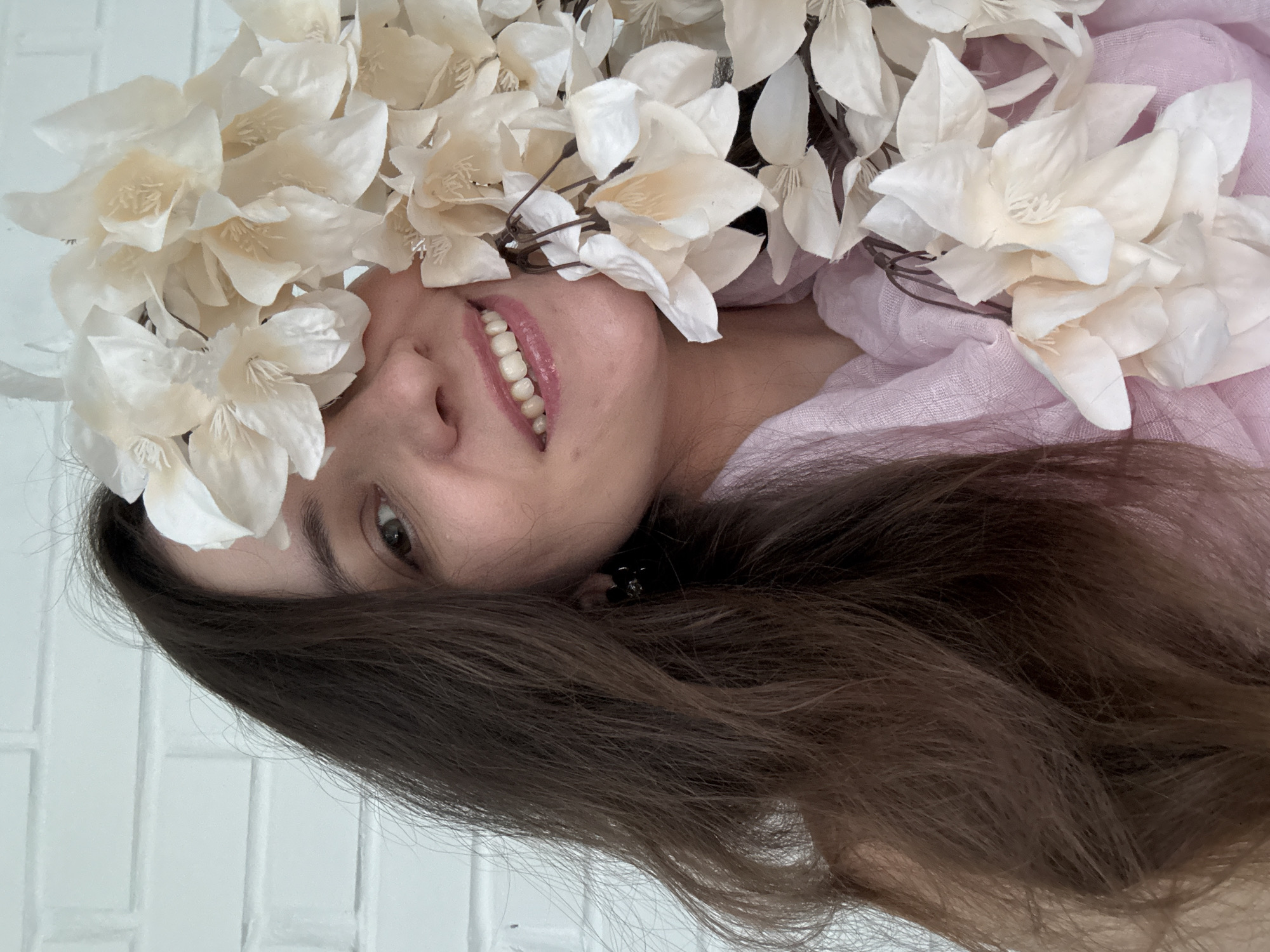 This close-up portrait features a young woman lying down, looking upwards with a gentle, pleasant smile that reveals her upper teeth. Her face is framed by a dense arrangement of white and cream-colored artificial flowers, which form a crown or headpiece covering parts of her forehead and cheeks. Her long, dark brown hair cascades around her head and shoulders.
She is wearing or lying on a soft, light pink or lavender fabric that is visible around her neck and to the side of her face. Her eyes are open and dark, looking directly at the viewer. The background behind her head and hair appears to be a light-colored wall, possibly with faint horizontal tile lines, suggesting an indoor setting. The lighting is soft and even, highlighting the textures of the flowers, hair, and fabric. The image is rotated 90 degrees clockwise, giving the impression of the subject lying on her back. There is no visible text.