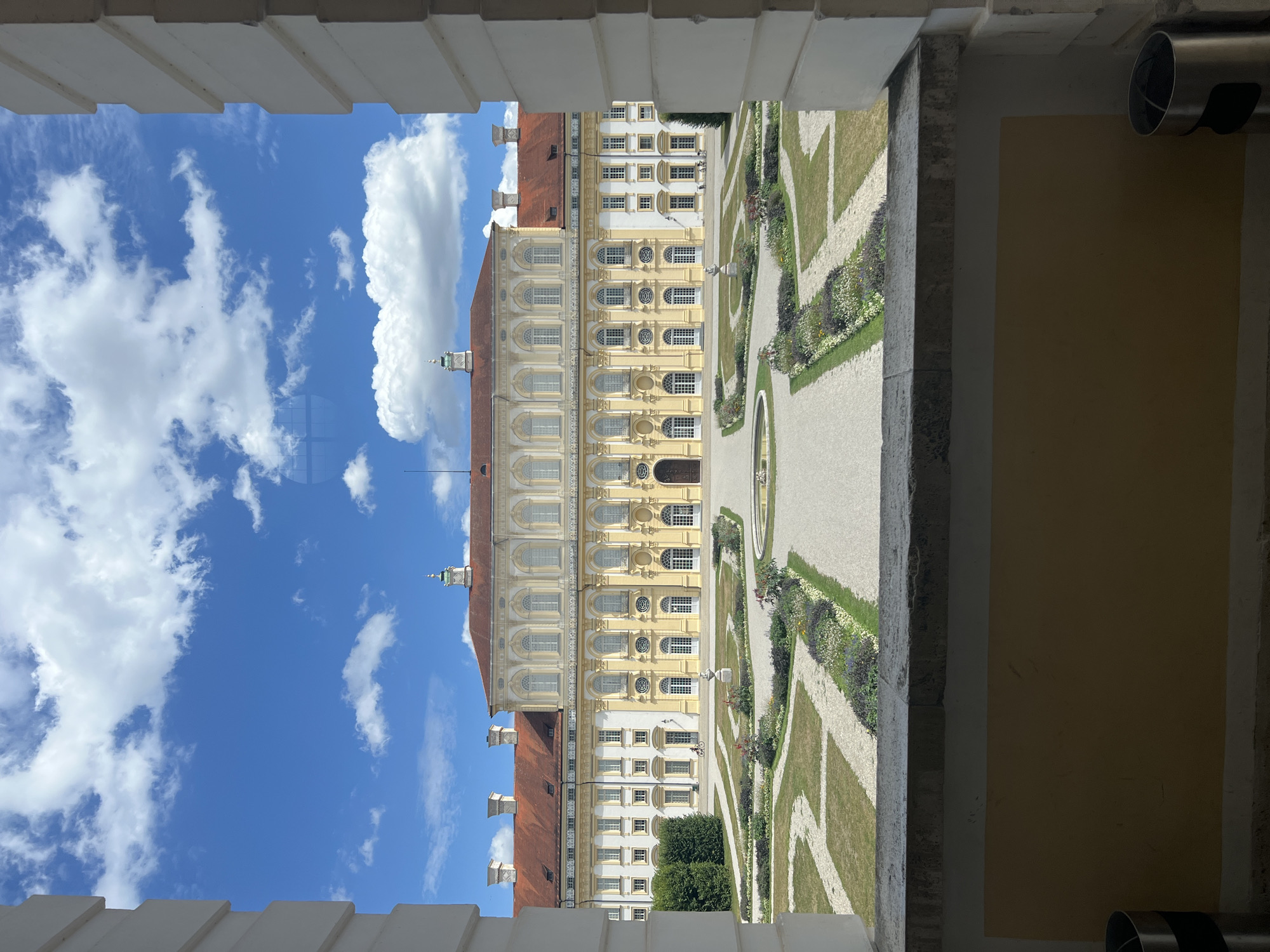 The image shows a Baroque palace and formal gardens viewed from an interior vantage point framed by architectural elements. The palace, painted in yellow and white with a red tile roof, features multiple stories, arched windows, and decorative stonework. In front of the palace lies a manicured garden with geometric pathways, flowerbeds, and a small fountain. The sky above is blue with scattered white clouds. To the left and right, the framing structure, likely part of a balcony or loggia, is visible, constructed of white stone. A person is seen on a bicycle in the middle ground, and other figures are visible in the distance on the palace grounds. The scene is set at Schleissheim Palace in Oberschleissheim, Germany.