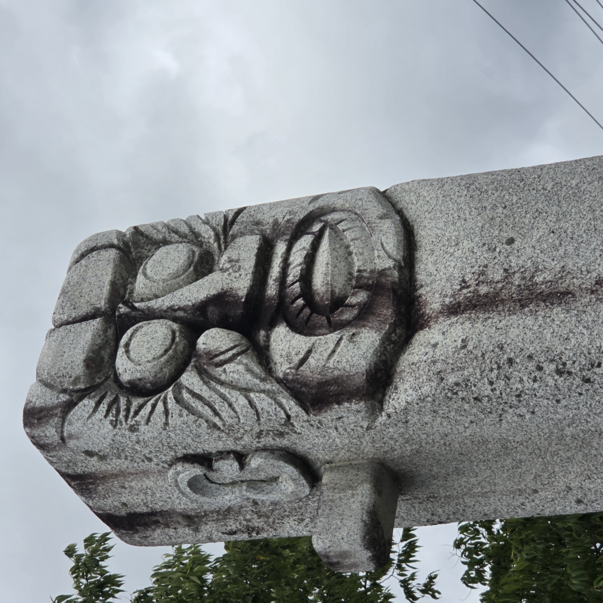 An outdoor, low-angle, medium shot captures a stone sculpture of a face, carved with exaggerated features such as bulging eyes, a wide mouth with teeth, and textured hair. The sculpture is made of speckled gray stone and shows dark staining on its surface, particularly around the facial features. To the left of the sculpture, green foliage from a tree is visible against a cloudy, overcast sky. The base of the sculpture extends downwards, out of view. The location is identified as Dunnae-myeon, South Korea. There are no people visible in the image.