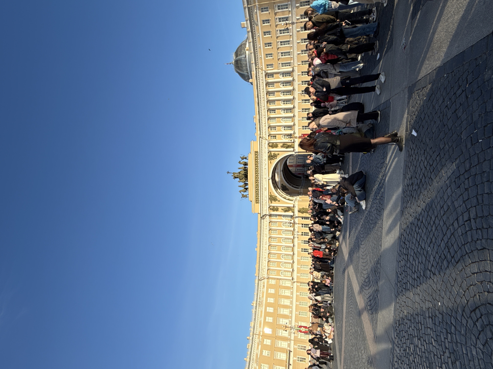A vertical photograph shows a large crowd of people assembled at the Palace Square in Saint Petersburg, Russia. The crowd is gathered near the General Staff Building, a long, yellow-colored edifice with a prominent arch topped by a sculptural chariot and horses. The sky above is clear blue. The ground is paved with cobblestones.