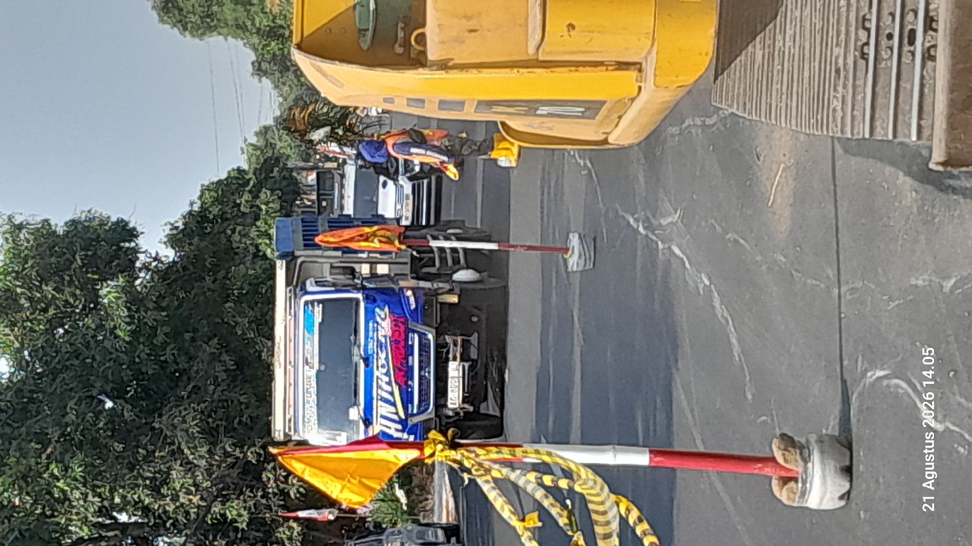 The image depicts a street scene in Takeran, Indonesia, on August 21, 2026, at 14:05. In the foreground, a construction site is evident, with a yellow excavator and yellow and black caution tape decorating red and white striped poles. The tape is strung across two poles, with one pole featuring a colorful orange flag on top, and the other pole, in the middle ground, also topped with an orange flag.

A blue dump truck with "HINTANG KENCANA" written on its side is parked in the middle of the road. The truck has a license plate that reads "AG 8999 VU". A person wearing a blue head covering, an orange safety vest, and yellow boots is standing near the excavator, seemingly supervising or working on the construction.

In the background, other vehicles are visible, including a white car, suggesting traffic is present or has been temporarily diverted. The environment appears to be a sunny day with trees lining the roadside and overhead power lines indicating an urban or semi-urban setting. The road surface shows signs of repair or ongoing construction, with white lines and patches visible.
