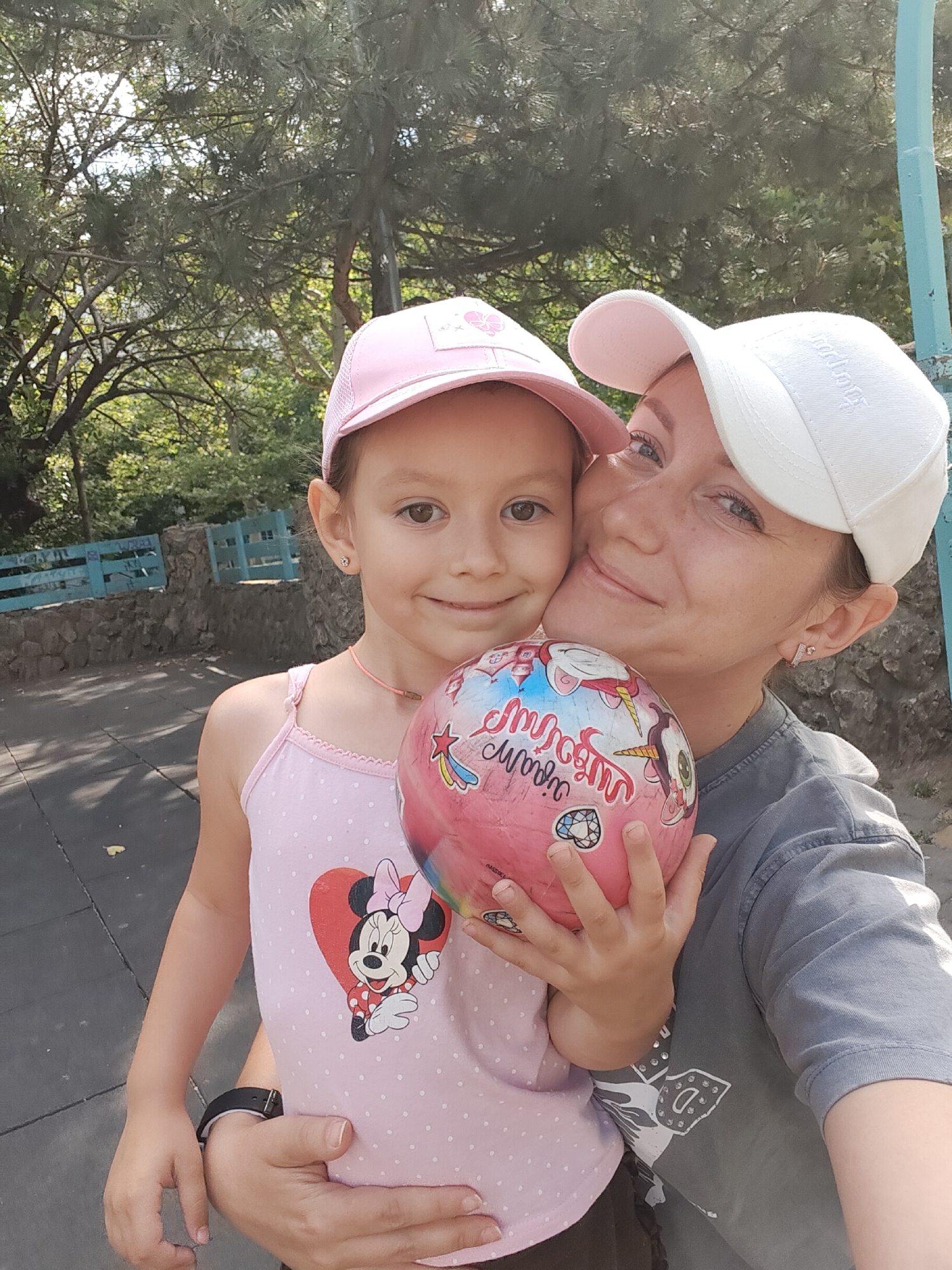 A selfie shows a woman and a young girl embracing outdoors. The girl, on the left, wears a pink hat and a pink tank top with a Minnie Mouse design. She holds a pink, multi-colored ball with cartoon characters. The woman, on the right, wears a white baseball cap and a gray t-shirt. Her arm is around the girl. They are in an outdoor area with trees and a stone wall in the background. The overall scene suggests a casual, happy moment between the two individuals.