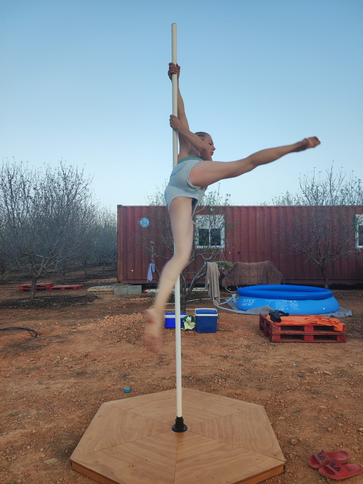 A person is performing a pole dance outdoors in Alcàsser, Spain. They are dressed in light blue shorts and are holding onto a white pole with their hands. Their body is arched backward, and one leg is extended outwards. The pole is mounted on a hexagonal wooden base. In the background, there is a large red shipping container with windows and a blue circular design. To the right of the container, there is a blue inflatable pool and a wooden pallet with orange fabric and a black bag on it. The ground is covered in reddish-brown dirt and small stones. There are trees with bare branches scattered throughout the scene. The sky is a clear light blue. Red flip-flops are visible on the ground in the lower right corner.
