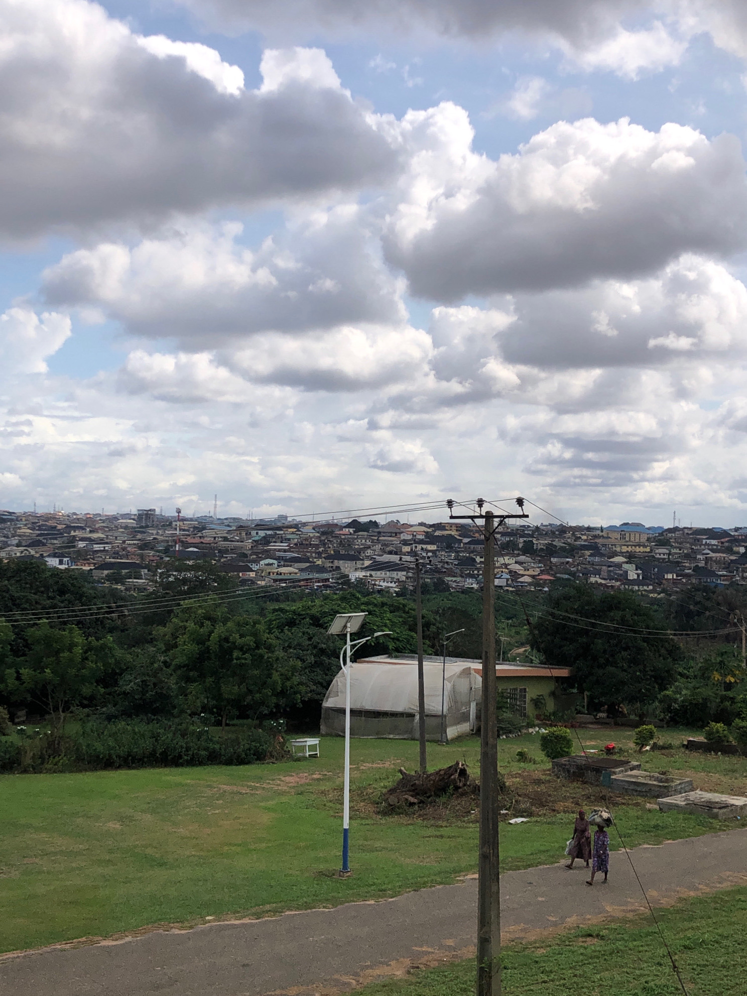 An aerial view shows a sprawling city in Lagos, Nigeria, under a cloudy sky. The city is densely packed with buildings of varying heights and colors, stretching as far as the eye can see. In the foreground, a lush green lawn slopes down to a paved pathway where two women are walking, one carrying a load on her head. A tall wooden utility pole with wires stands to the right, and a greenhouse is visible in the mid-ground. The sky is filled with dramatic, grey and white clouds, suggesting an overcast or potentially rainy day. The image captures a vibrant urban landscape contrasted with the natural elements of the foreground and the sky.