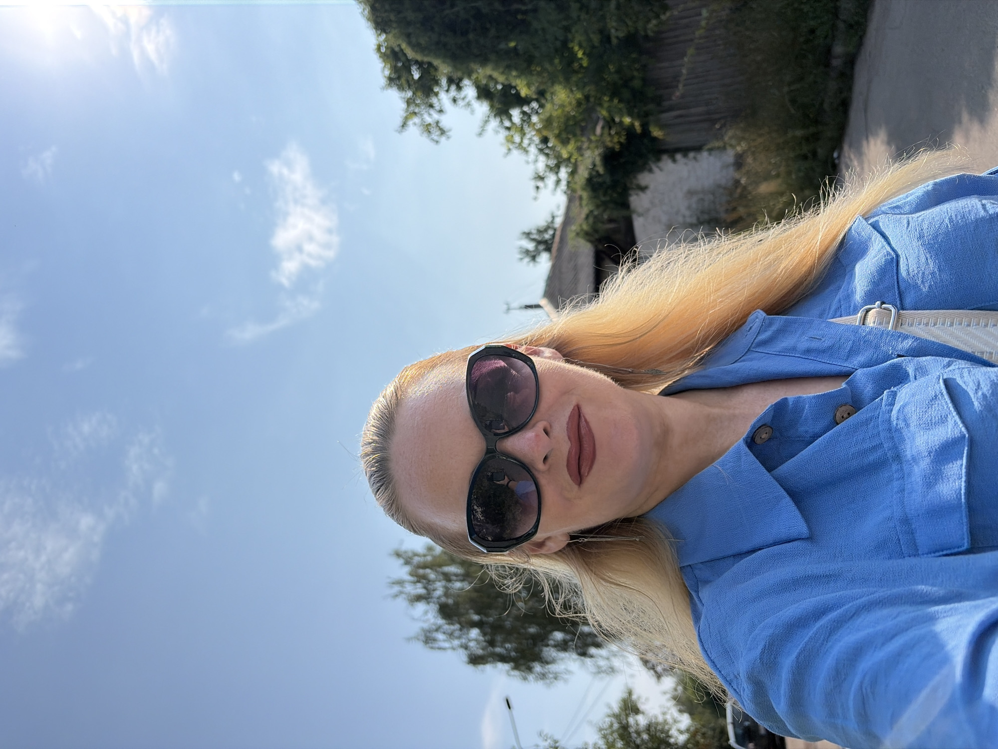A woman with blonde hair is taking a selfie outdoors. She is wearing a blue collared shirt with buttons and pockets, dark sunglasses, and has on dark lipstick. Her hair is long and wavy, and she has on silver earrings. In the background, there are trees, buildings, a fence, and a road. The sky is bright blue with some white clouds. The sun appears to be shining.