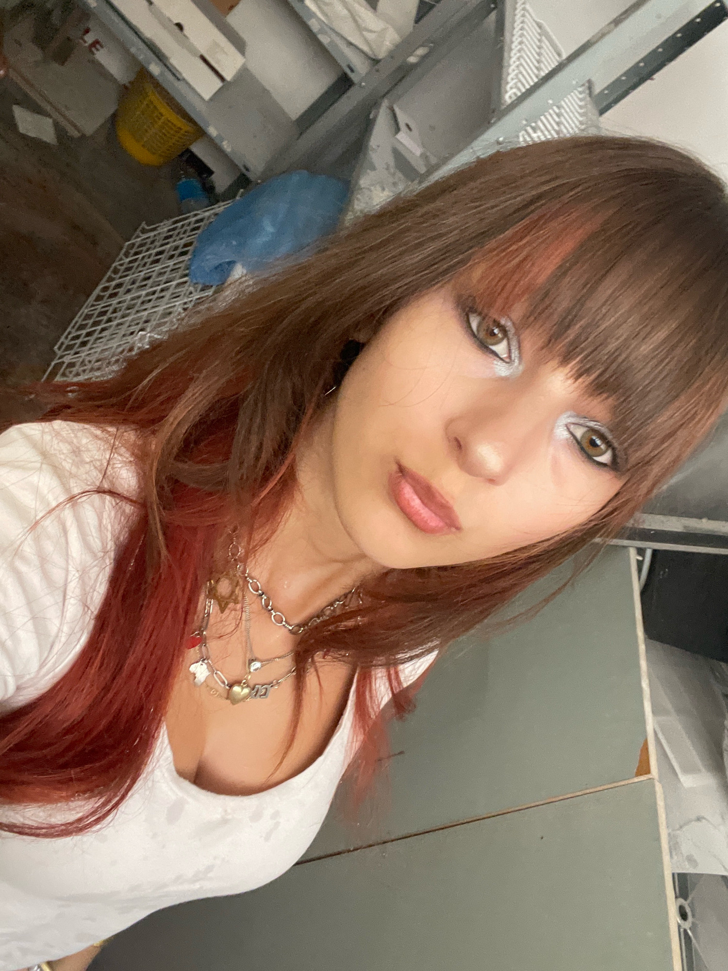 A young woman with brown hair featuring reddish highlights and bangs is looking at the camera. She is wearing a white v-neck top and multiple silver necklaces, one with a gold heart charm. Her makeup includes silver eyeshadow and dark eyeliner. The background appears to be an indoor area with various objects, including a yellow basket, a blue plastic bag, and a white metal rack. Some shelving units are visible, and the overall environment seems somewhat cluttered or unfinished. There is no identifiable context or details that would place this scene in Raanana, Israel.