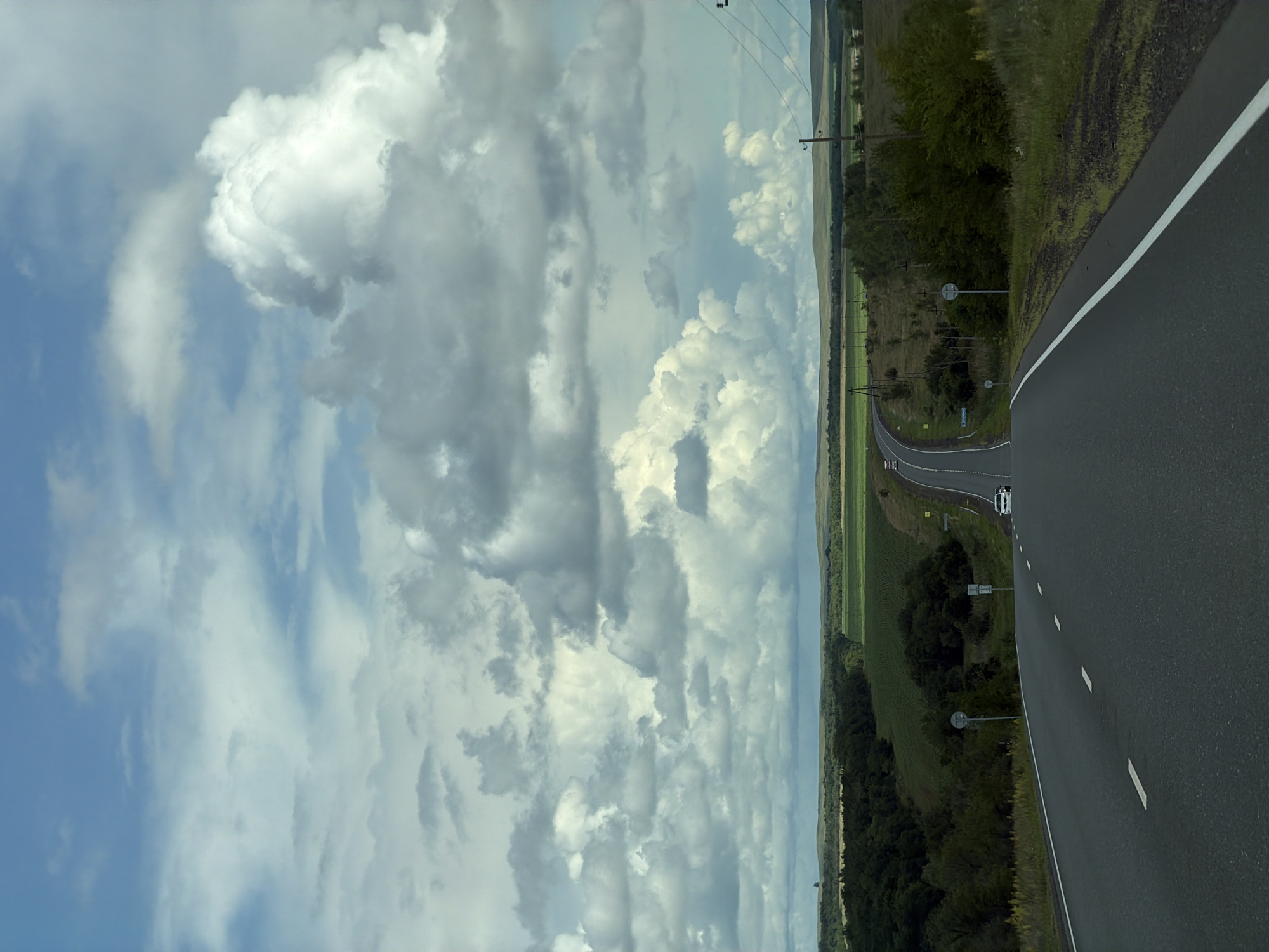 The image depicts a view from a car on a paved road leading through a rural landscape under a dramatic, cloudy sky. The road curves to the right and then ascends a gentle hill, with white lane markings visible. On either side of the road, there is green vegetation, including trees and fields. In the distance, rolling hills are visible under a sky filled with large, puffy white and gray clouds. Two white cars and a red truck are seen on the road in the distance. Electrical poles and wires run along the right side of the road. The scene appears to be set in a natural, undeveloped area.