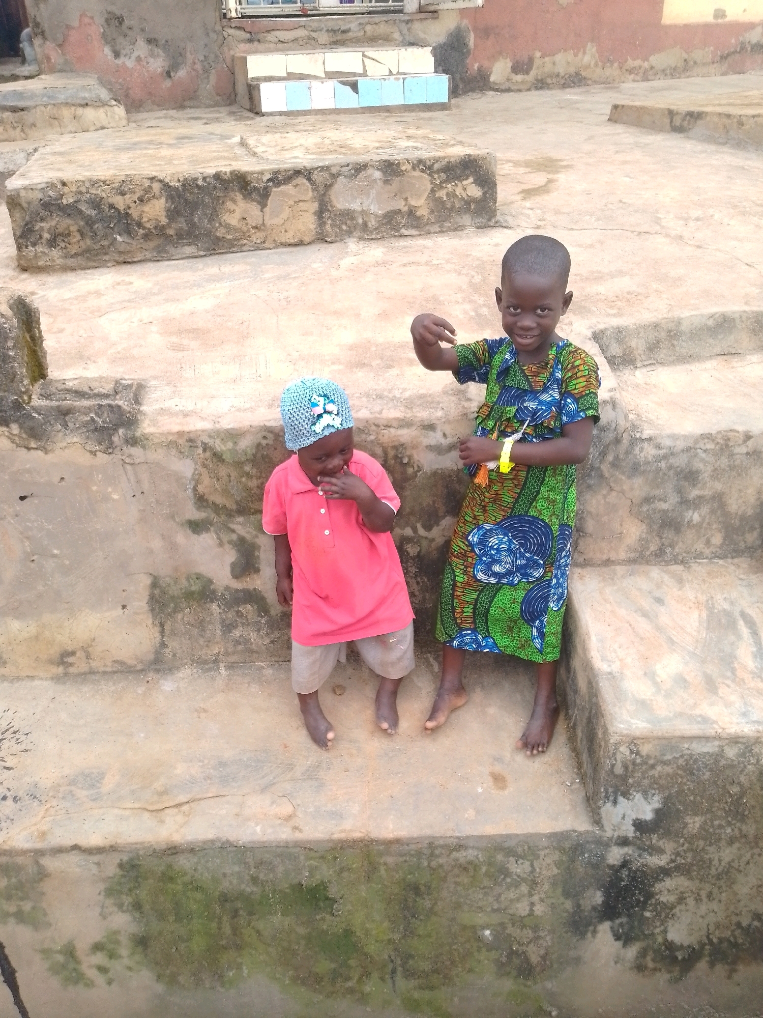 Two young children, a boy and a girl, are standing on concrete steps in an outdoor setting, possibly in Lagos, Nigeria. The boy, on the right, is wearing a vibrantly colored green and blue patterned dress and has his right hand raised. The girl, on the left, is wearing a pink t-shirt, light-colored shorts, and a light blue knitted hat with white embellishments. She has her left hand to her mouth. The steps are worn and appear to be made of concrete, with some patches of moss and discoloration. The background shows a building with weathered walls.