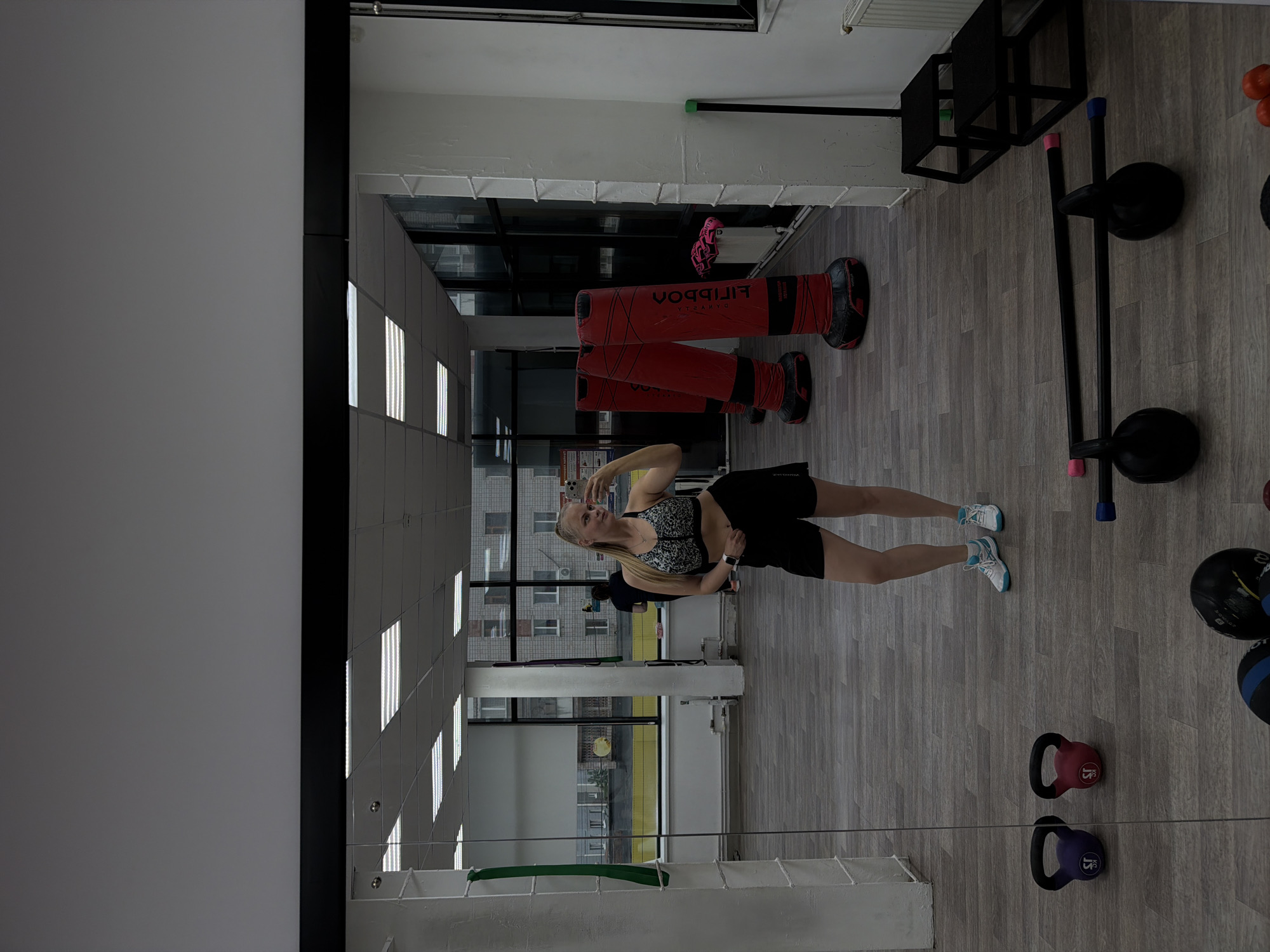 A woman is taking a selfie in a gym. The gym contains punching bags, weights, and workout equipment. The gym appears to be on the second floor of a building in Izhevsk, Russia. A residential building is visible through the windows.