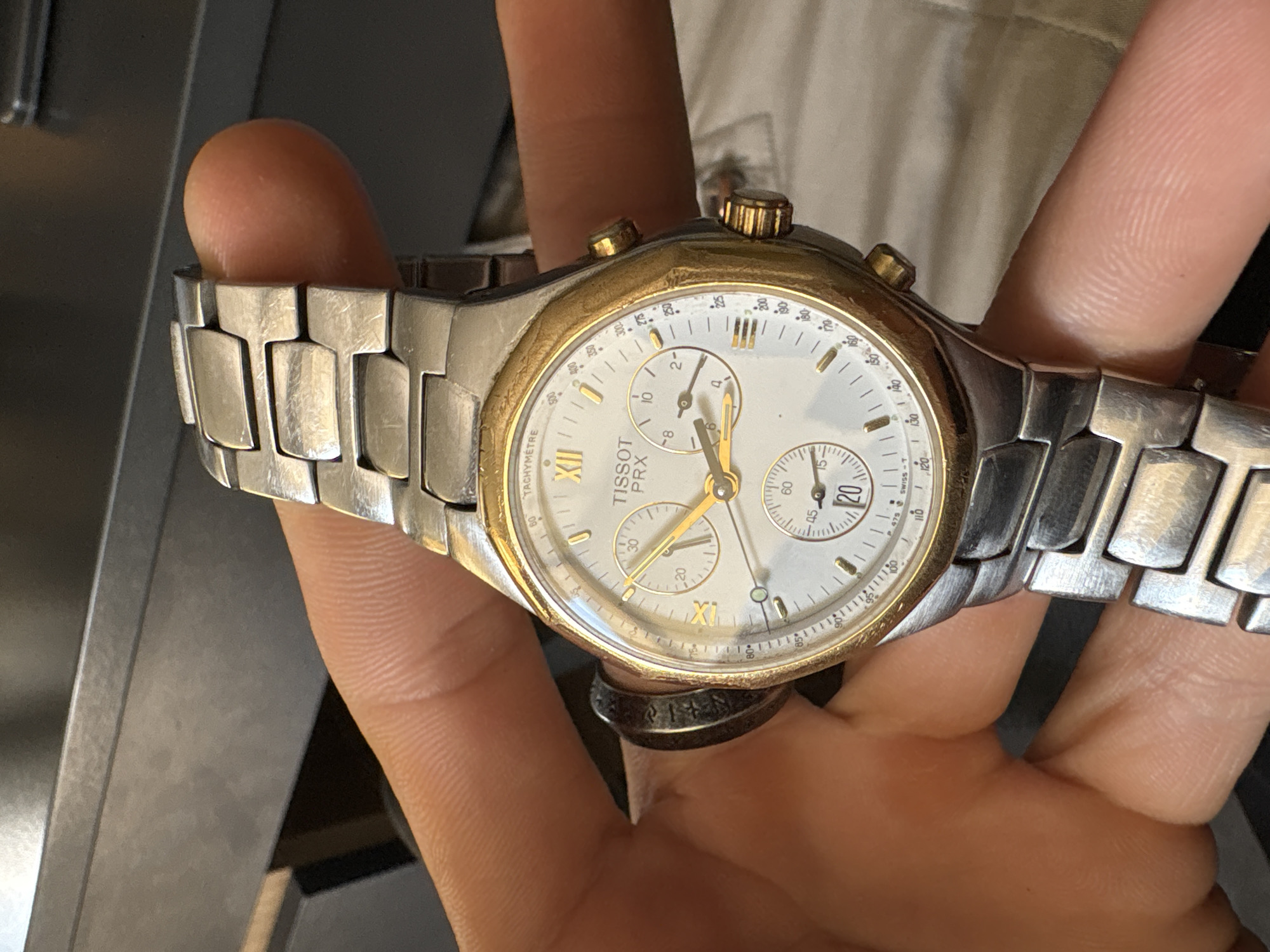 A hand holds a Tissot PRX chronograph watch. The watch has a white dial with gold accents, a stainless steel band, and gold-colored pushers. The dial features chronograph subdials and a tachymeter scale around the edge. The watch is being held against a background that includes a piece of light beige cloth and a dark grey surface, possibly a table or cabinet. A dark ring is visible on the ring finger of the hand holding the watch. The image is well-lit and in focus. There is no indication of the image being taken in Sofia, Bulgaria.