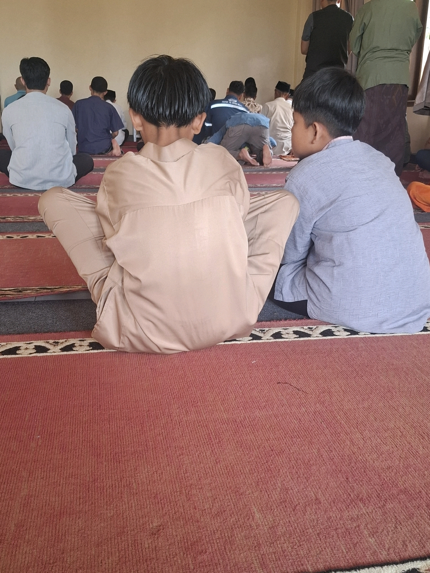The image shows a group of people sitting on a red carpet in a room, likely a mosque in Ciheulang Tonggoh, Indonesia, during a prayer service. The perspective is from behind, focusing on the backs of two young boys in the foreground, dressed in tan and light blue traditional clothing. Other individuals, mostly men, are visible further back, some kneeling in prayer, others sitting. The room has a light-colored wall and a patterned carpet.