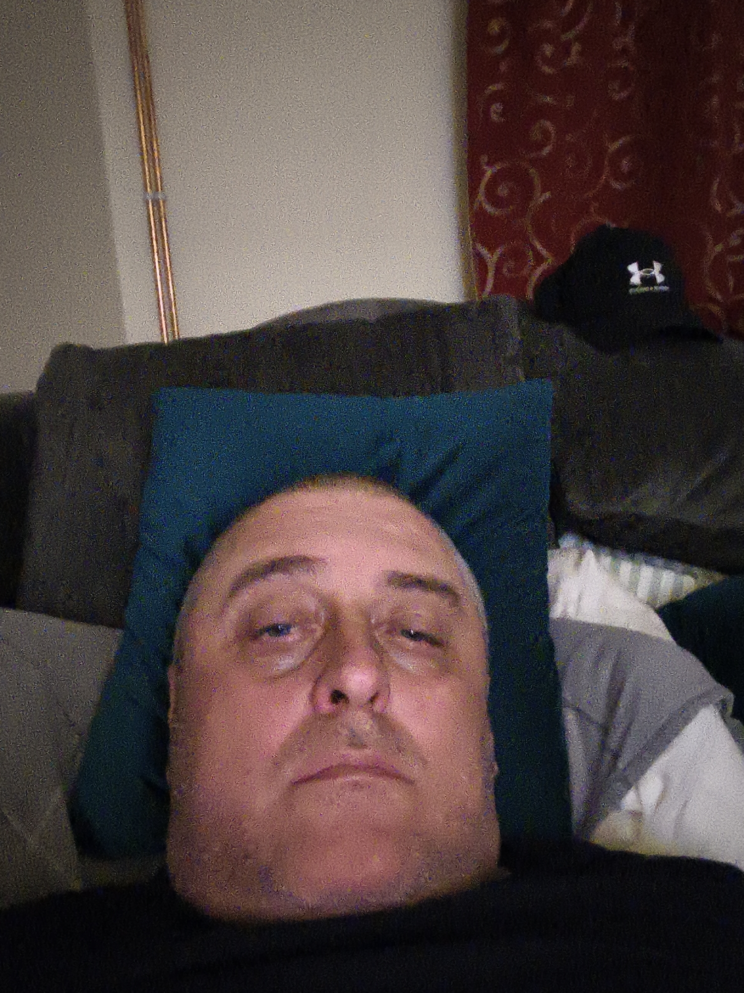 A man is depicted resting indoors, likely in a living room or bedroom in Huntingdonshire, United Kingdom. He is seen from the chest up, lying down with his head on a dark teal pillow. He has fair skin, a receding hairline, and light stubble on his face, wearing a dark-colored shirt. His expression is neutral, appearing relaxed or slightly tired, as he looks directly towards the camera.
In the background, to the man's left, a dark grey couch or sofa is visible. On the right side of the frame, a black baseball cap with a white "Under Armour" logo rests on the couch, positioned in front of a red curtain with a circular pattern. Two thin, vertical, copper-colored pipes are visible against a light-colored wall in the upper left portion of the image. Part of a white and grey striped pillow or bedding is also visible on the far right, beside the man. The lighting appears dim, and the image exhibits noticeable digital noise or grain, suggesting it was taken in low light conditions, possibly during the evening or night.