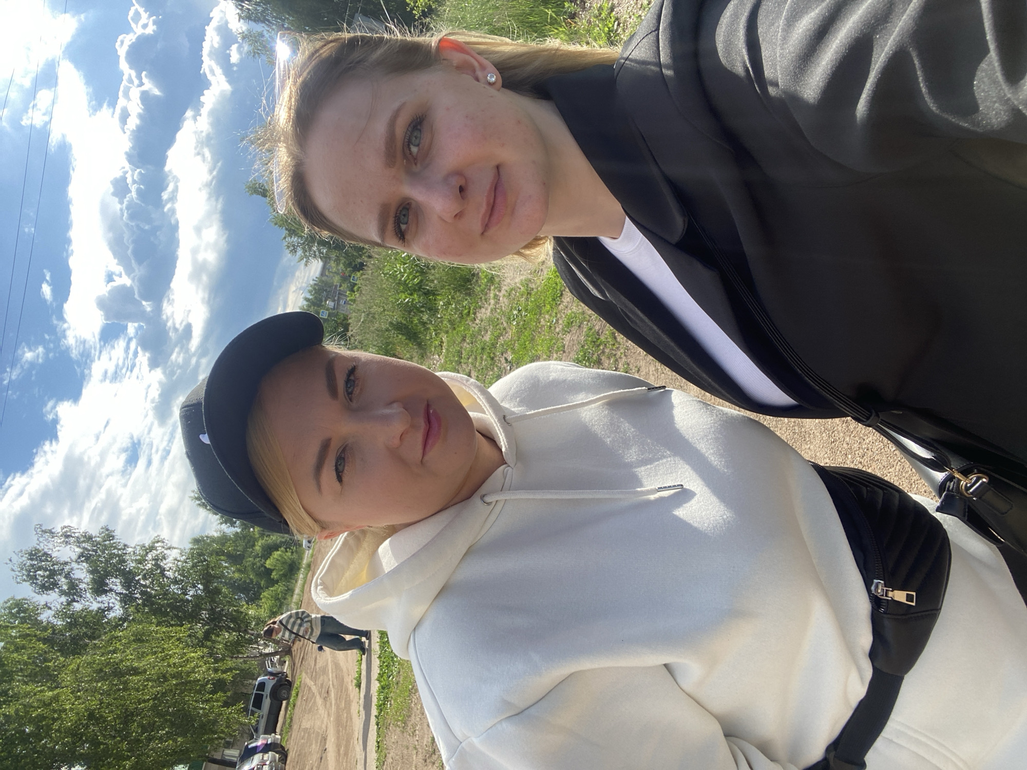 Two women are standing outdoors, posing for a selfie. The woman on the left is wearing a white hoodie, a black baseball cap, and a black fanny pack. The woman on the right is wearing a black blazer over a white shirt and a small stud earring. In the background, there are trees, cars, and a cloudy sky. The scene appears to be a street in the city of Chaikovsky, Russia.