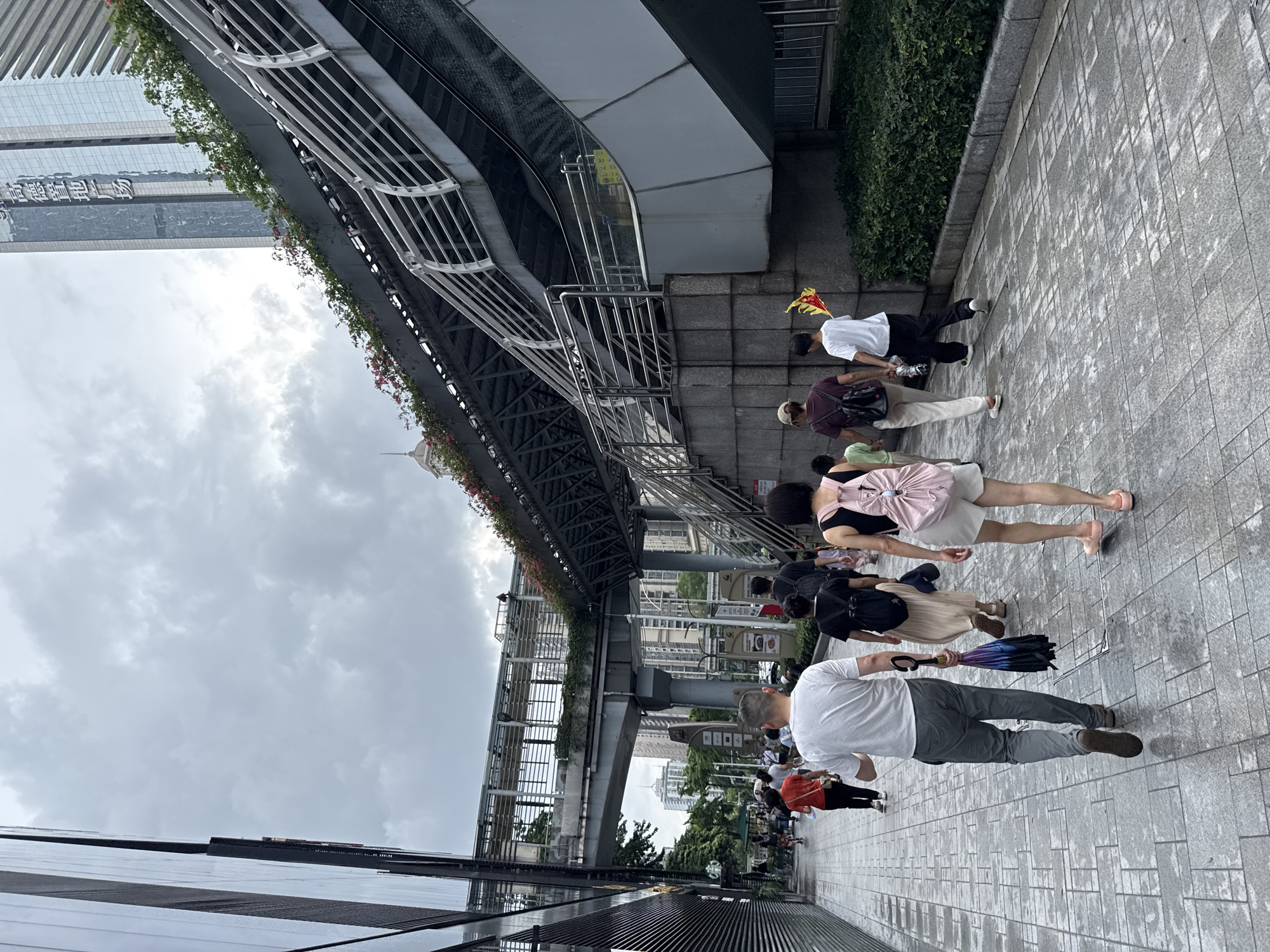 A medium shot captures a group of people walking on a wet, tiled sidewalk in Tianhe District, China, under a cloudy sky. The perspective is from behind the walking individuals. To the left, a modern building with a glass facade and dark horizontal accents dominates the scene. Ahead, an elevated pedestrian walkway, supported by gray pillars, stretches across the frame, featuring metal railings and planters with red and pink flowers. To the right, a multi-level structure with gray concrete and metal railings, possibly an escalator or staircase, is visible, also adorned with greenery. In the background, tall modern buildings with glass exteriors rise into the overcast sky.
The people in the foreground are walking away from the camera. Closest to the camera is a man in a white t-shirt and gray pants, carrying a folded umbrella with a patterned fabric. Next to him, a woman wears a pink backpack, white shorts, and pink sandals. Behind them, a person in a dark shirt and beige skirt walks with a black backpack. Further ahead, a man in a white t-shirt and black pants is seen, holding a yellow and red flag or kite. Another individual in a dark t-shirt and beige pants walks alongside him. Several other people are visible walking in the distance along the sidewalk and on the elevated walkway. The wet pavement suggests recent rain.