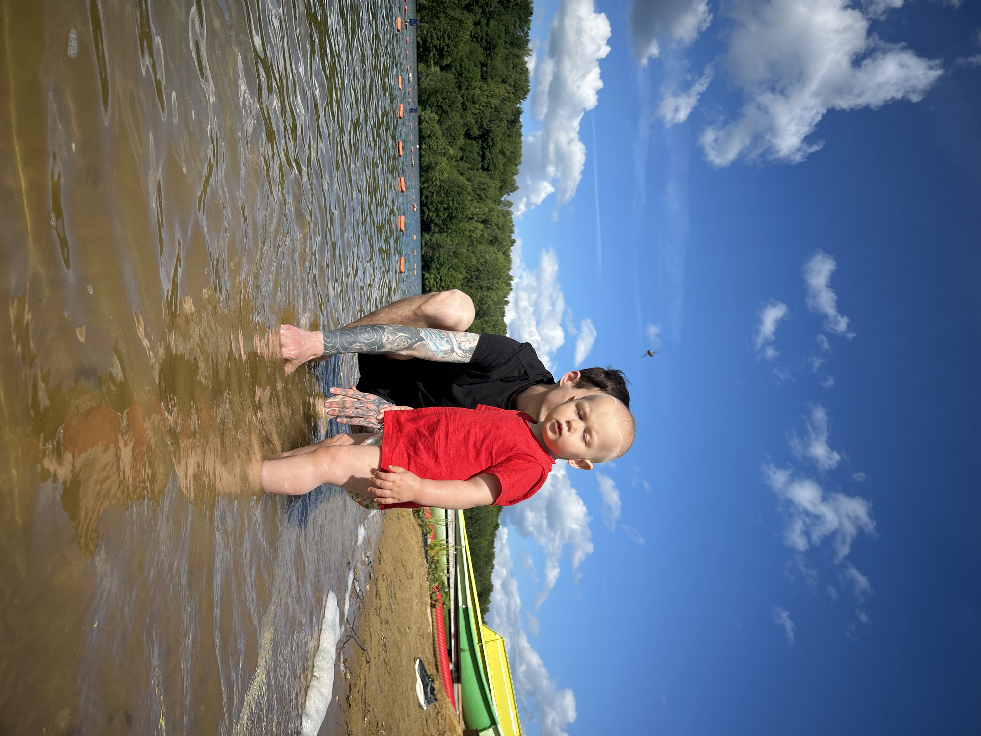 A man in a black t-shirt with tattoos on his arm is crouching in shallow water next to a toddler wearing a red t-shirt and patterned shorts. They are at the edge of a lake with a sandy beach and green trees in the background. Several colorful boats are visible on the beach. The sky is blue with white clouds.