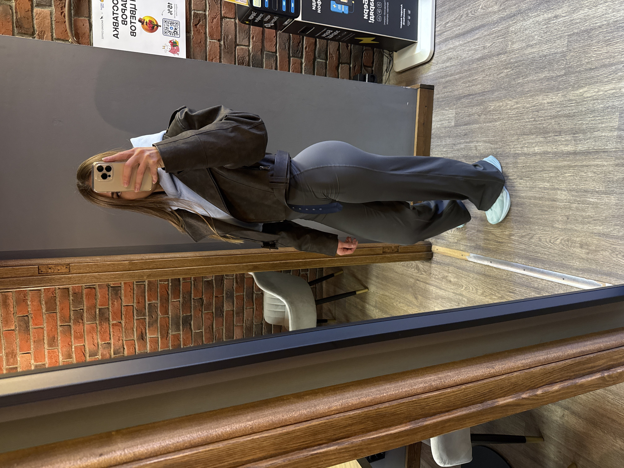 The image appears to be a selfie taken in a mirror. The subject is a person with long brown hair holding a smartphone in their left hand, presumably taking the photo. They are wearing a brown jacket over what appears to be a light-colored hoodie, and gray leggings or pants. They are wearing light blue sneakers.
The background visible through the mirror and surrounding the subject includes a gray wall, a brick wall (likely a reflection), and a section of wooden flooring. There's also a charging station or device with various phone charging cables visible. The selfie is taken in Saint Petersburg, Russia, and also visible is a white chair reflected in the mirror.