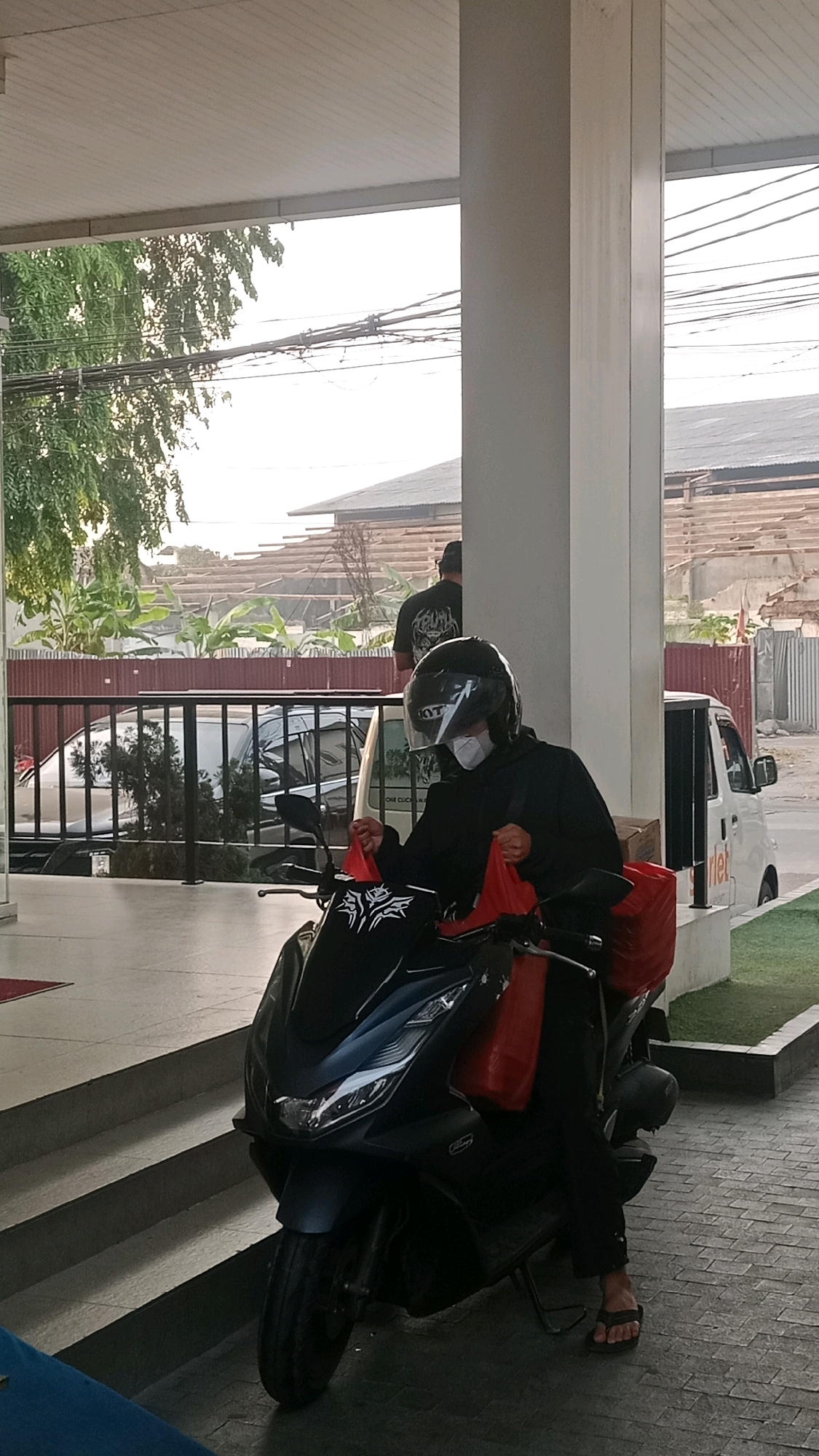 In West Jakarta, Indonesia, a person wearing a black hoodie, black helmet, and a white face mask is seated on a black scooter. They are holding two red bags, one on each side of the handlebars. The scooter is parked on a paved area next to a set of concrete stairs. Behind the scooter, a white van and a dark-colored car are visible. In the background, a building under construction with exposed wooden beams and a corrugated metal roof is seen, along with trees and power lines. The overall scene appears to be an outdoor area of a commercial or residential building.