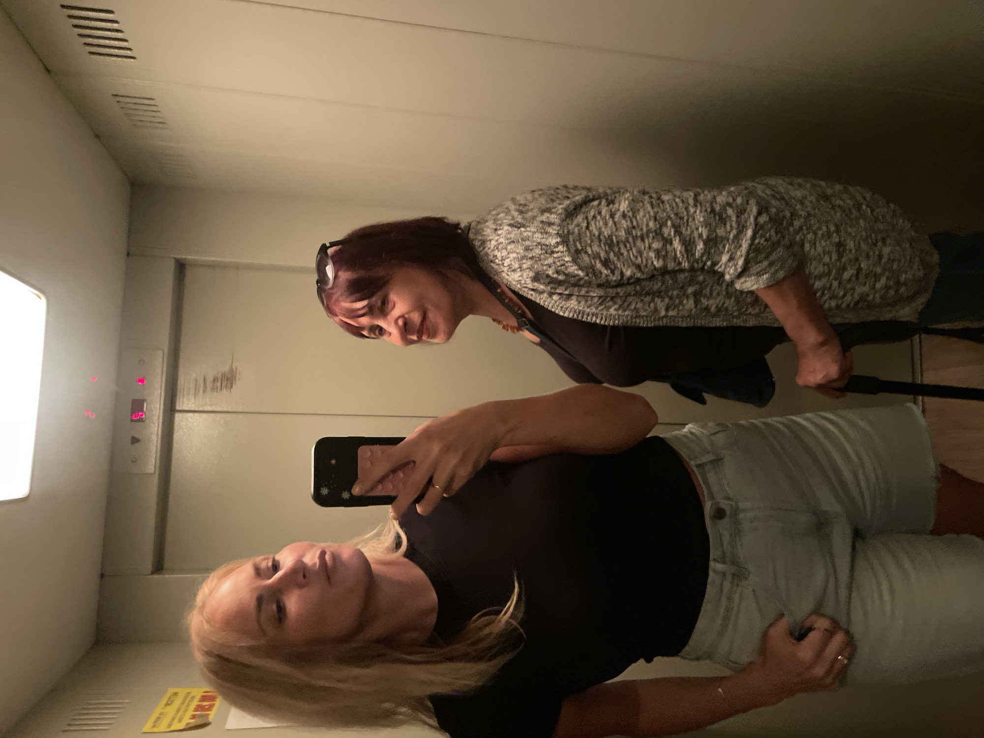 Two women are in an elevator. One is blonde and fair-skinned, wearing a black shirt and light-wash denim shorts, and is holding a black smartphone, taking a selfie in the mirror. The other woman has dark hair and is wearing a gray and white cardigan over a black top. The elevator has a panel with numbered buttons, and the display shows the number "5". The elevator walls are a light neutral color. The location of the photo is the city of Sochi, Russia.