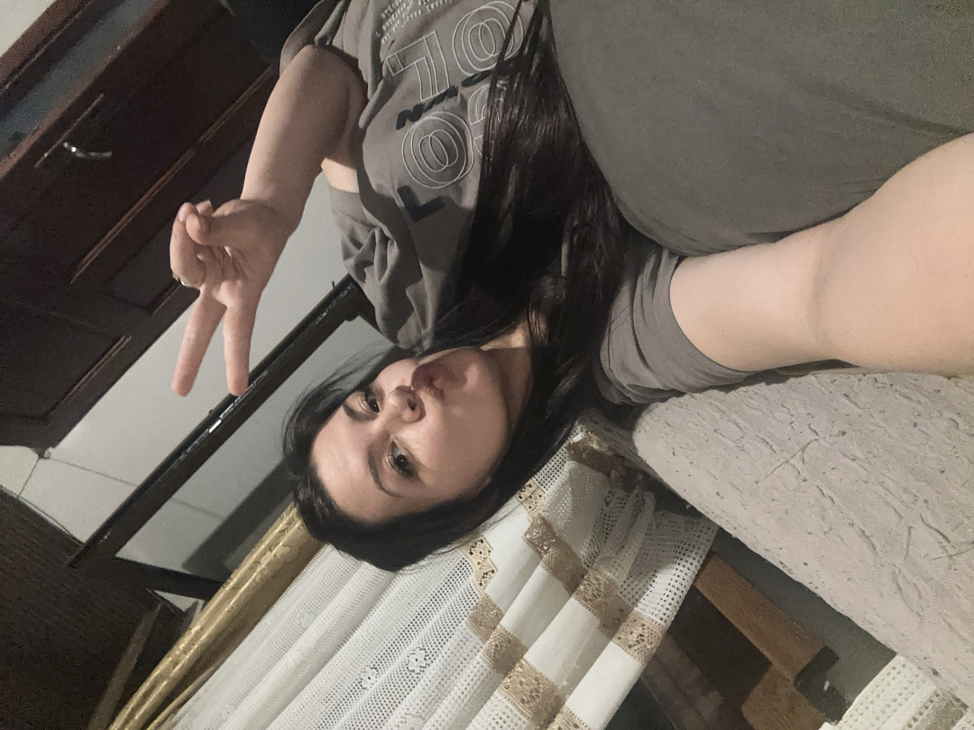A young woman is captured in a rotated, indoor selfie, likely taken in Chubra, Bulgaria. She is lying on her side, looking directly at the camera with a playful expression, her lips slightly puckered. Her long, dark hair is draped around her head. She wears a grey t-shirt featuring a graphic design of white outlined circles and stylized text, which appears to include "SOL" or "LOO," along with smaller, less legible black text.
Her right arm is raised, forming a peace sign (V-sign) with her index and middle fingers. The background shows various indoor elements: a dark wooden door with a silver handle to the left. To her right, a portion of a grey, textured surface, possibly a sofa or bed, is visible. Below her, a white, intricately patterned tablecloth with gold-colored woven designs covers a surface, suggesting a table. The lighting is soft, indicative of an indoor setting, possibly during the evening. The overall mood is casual and lighthearted.