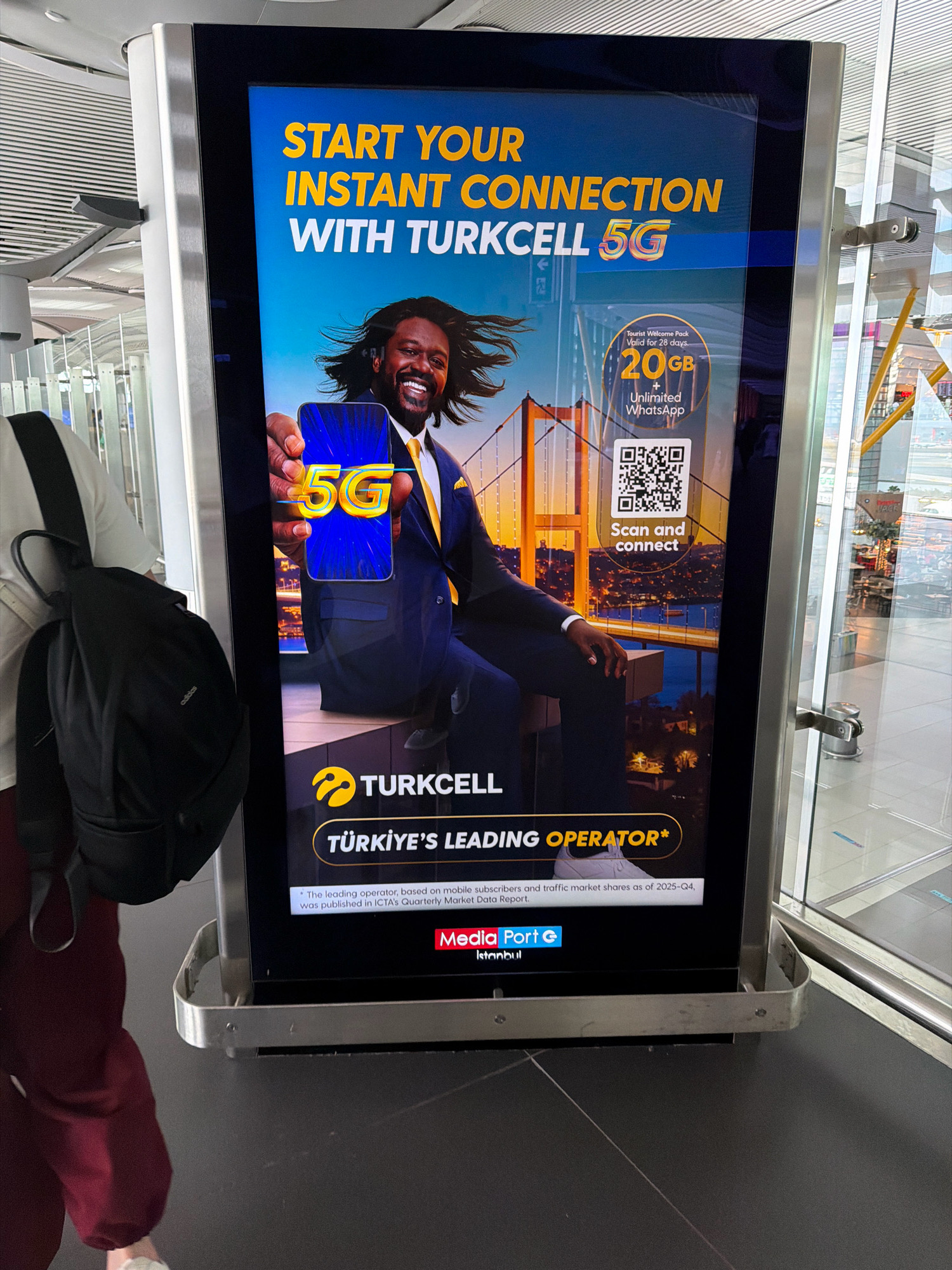 An advertisement for Turkcell 5G is displayed on a large digital screen at an airport, likely in Arnavutköy, Turkey, given the advertisement's content. The screen features a man in a suit and tie holding a smartphone displaying "5G." Text above the man reads "START YOUR INSTANT CONNECTION WITH TURKCELL 5G." A graphic indicates a "Tourist Welcome Pack" with "20GB" and "Unlimited WhatsApp." Below the graphic is a QR code with the text "Scan and connect." The bottom of the ad states "TURKCELL" and "TÜRKİYE'S LEADING OPERATOR*." A person carrying a black backpack on their left side, wearing a white shirt and maroon pants, is partially visible on the left edge of the frame, walking away from the camera. The background shows airport architecture with glass walls and structures.