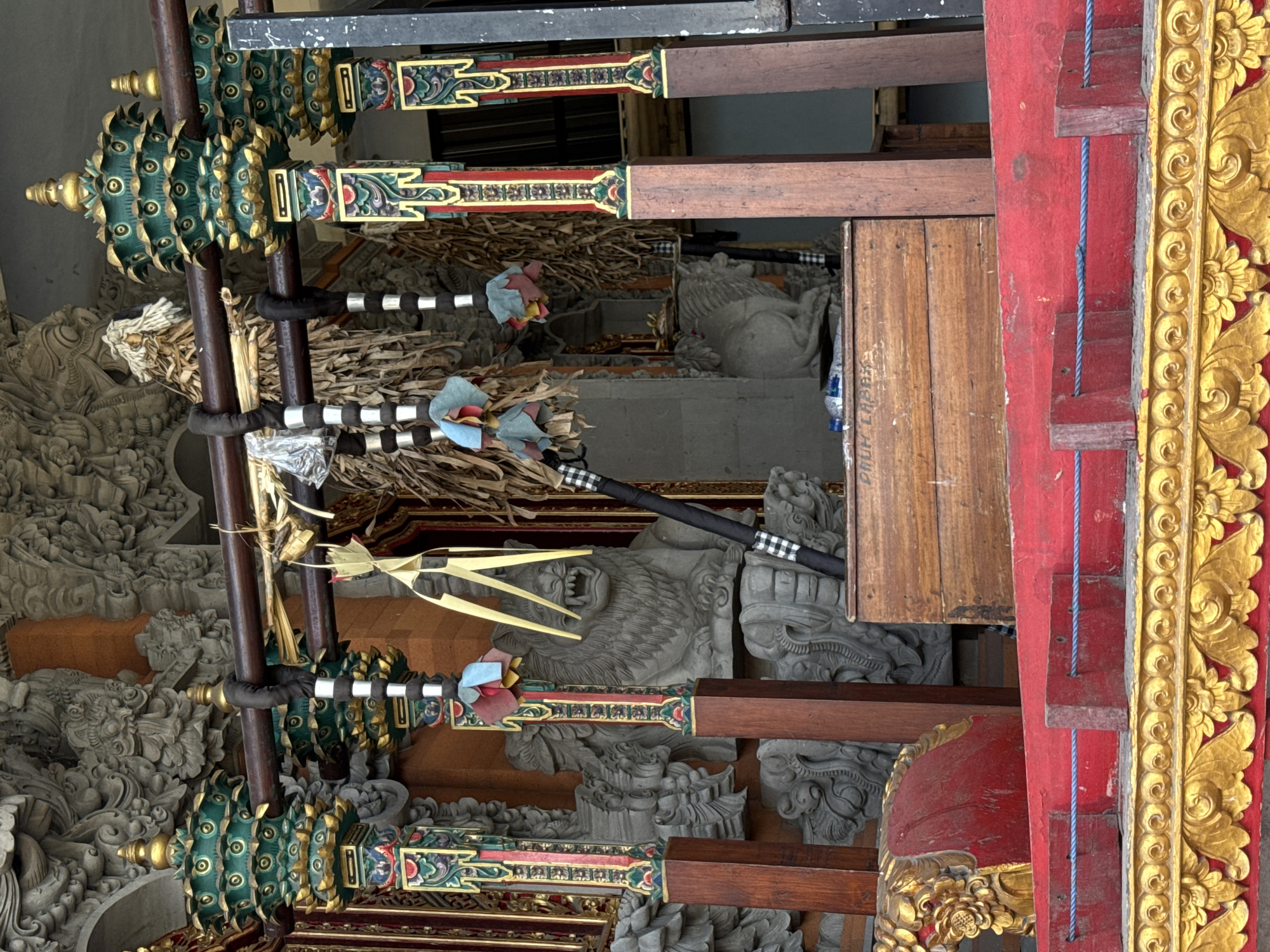 The image depicts a detail of a traditional Balinese gamelan instrument set against a backdrop of intricate stone carvings, likely located in a temple or cultural complex in Ubud, Indonesia. In the foreground, a vibrantly red and gold ornate wooden structure is visible. Above this, a wooden box rests on a dark surface. Suspended from horizontal wooden beams are various decorative elements, including dried palm leaves, tassels in muted colors of red, blue, and yellow, and what appear to be small, folded paper or fabric ornaments. Black fabric wraps around some of the hanging elements, and a black object with a white checkered pattern leans against the wooden box. In the background, large stone sculptures of what appear to be lions or mythical beasts are prominently displayed, integrated with elaborately carved architectural features. The overall scene suggests a cultural or ceremonial setting, rich in traditional Balinese art and symbolism.