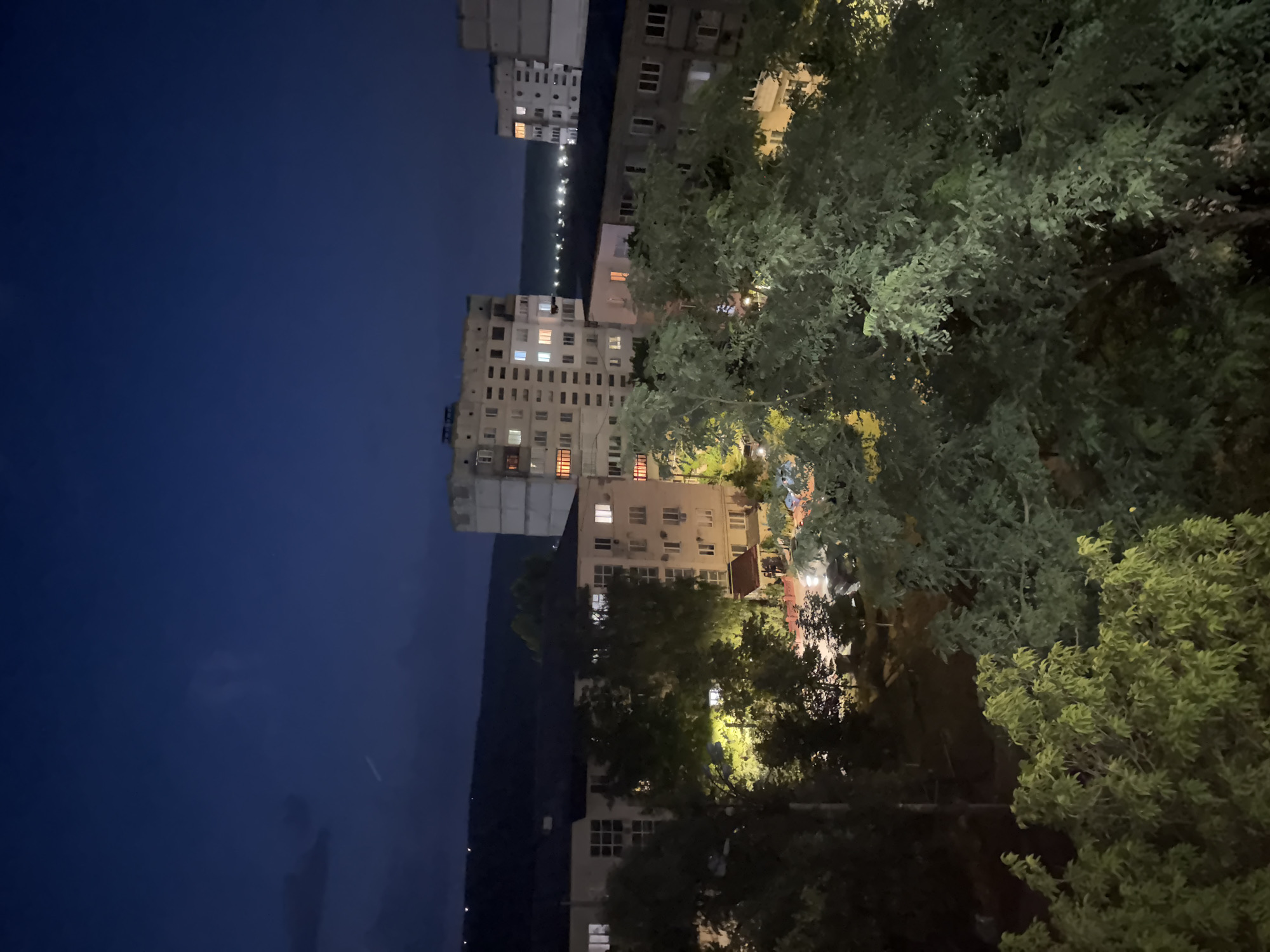 The image depicts a nighttime view of an urban landscape in Strășeni, Moldova. Several apartment buildings of varying heights are visible, with some windows illuminated, indicating occupancy. In the foreground, lush green trees obscure parts of the buildings, suggesting a residential area with significant vegetation. The sky is dark blue, transitioning to a lighter shade near the horizon, where a line of distant lights suggests a more densely populated area or road. The overall impression is one of a quiet evening in a city with both modern architecture and natural elements.