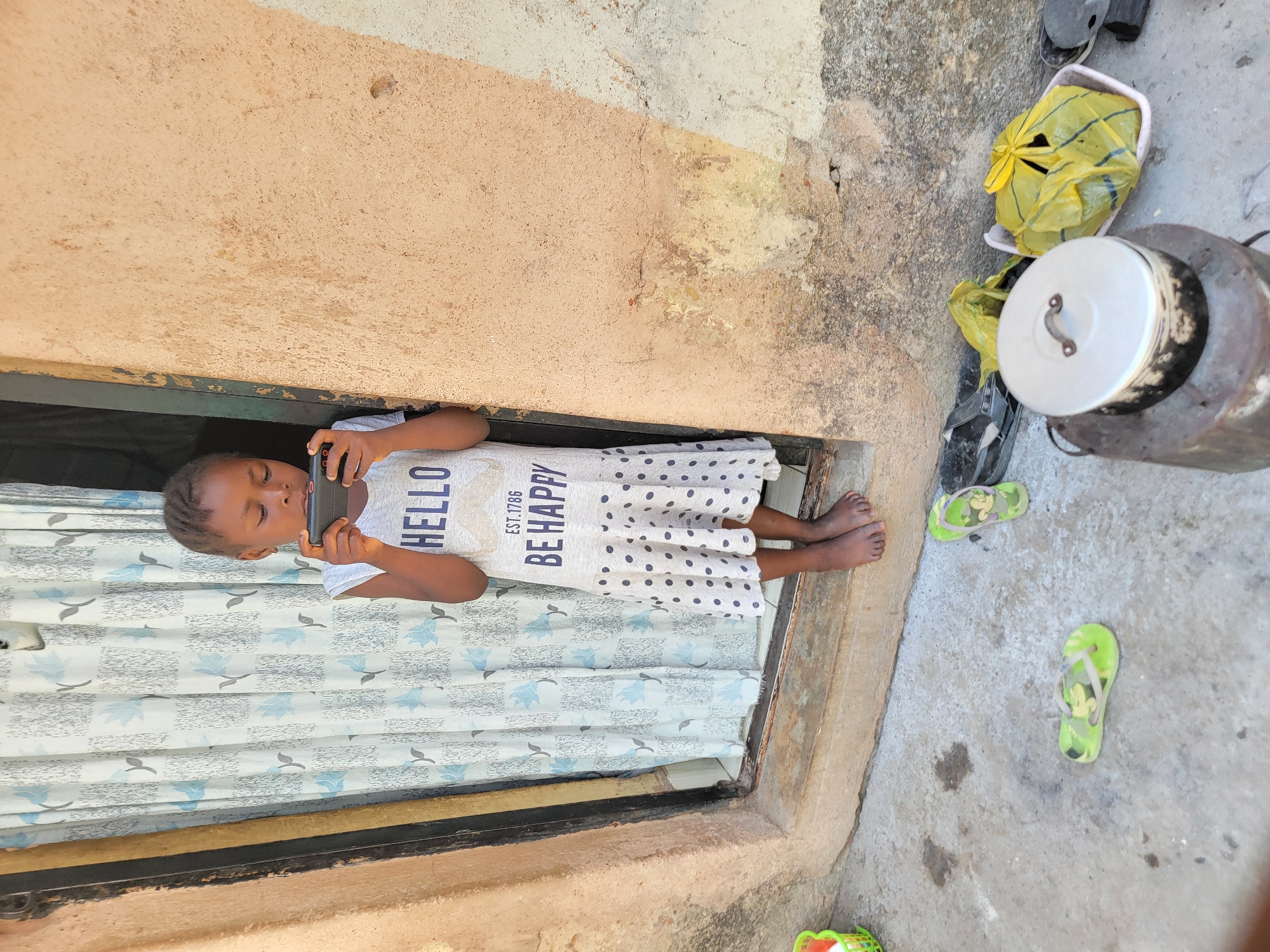 A young girl stands in a doorway, focused on a smartphone she holds horizontally in front of her. The girl is wearing a light-colored dress with blue polka dots. The dress has "HELLO" and "BE HAPPY EST. 1786" printed on it. A curtain with a blue and white floral pattern hangs behind her. The doorway is framed by a wall painted in a faded peach color on one side and bare concrete on the other. Outside the doorway, the ground is concrete and contains a yellow trash bag, green sandals, and a cooking pot. The location is likely in Jos, Nigeria, based on the context of the request.