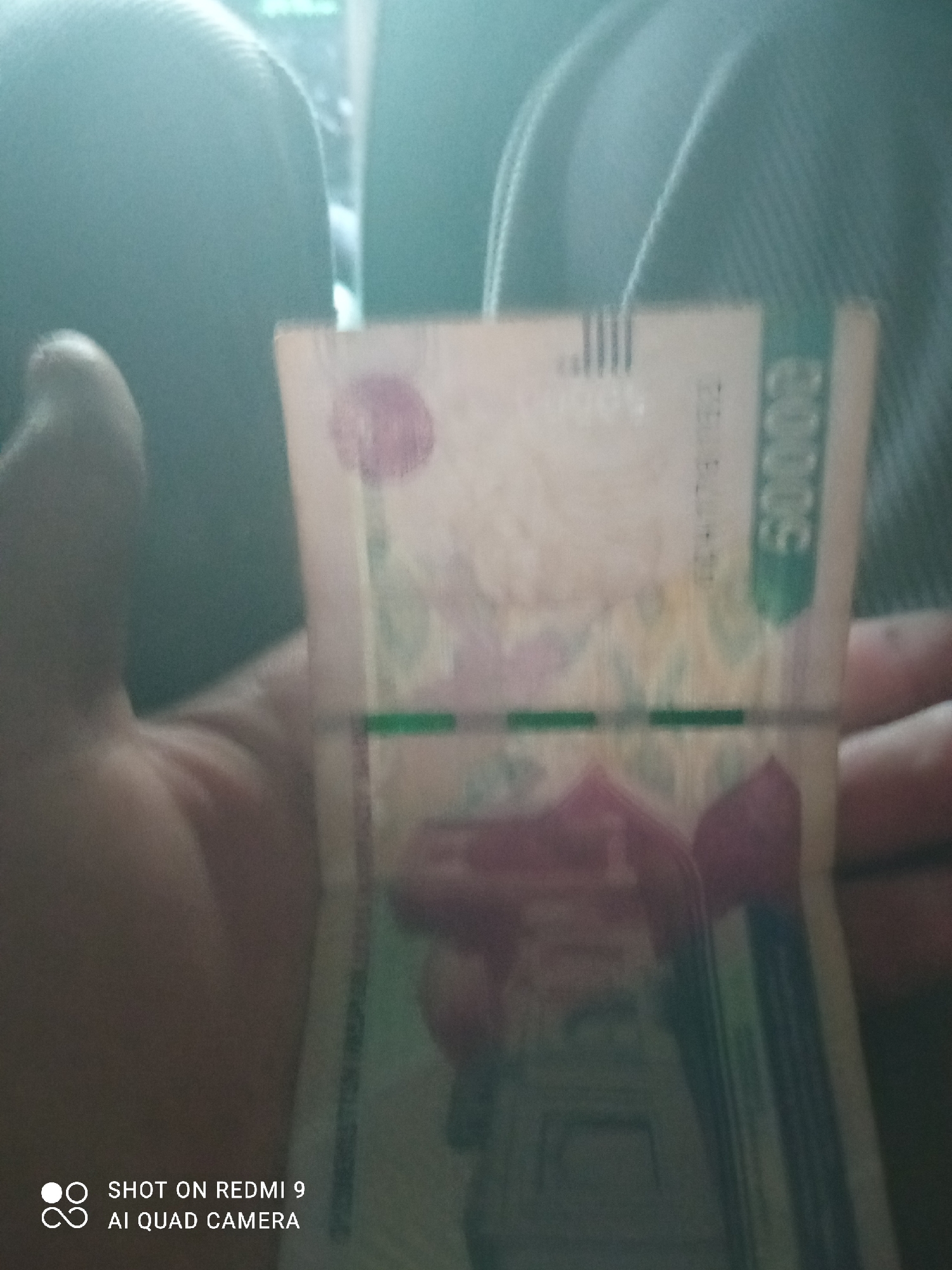 A hand is holding a banknote, possibly in the city of Gurlan, Uzbekistan. The banknote is partially visible, showing the number "5000" in green text on the right side. There are also pink and green markings on the banknote. The background is blurry, but appears to be an indoor setting with a dark, textured surface, possibly clothing or upholstery. The bottom left corner of the image shows a watermark with the text "SHOT ON REDMI 9 AI QUAD CAMERA."