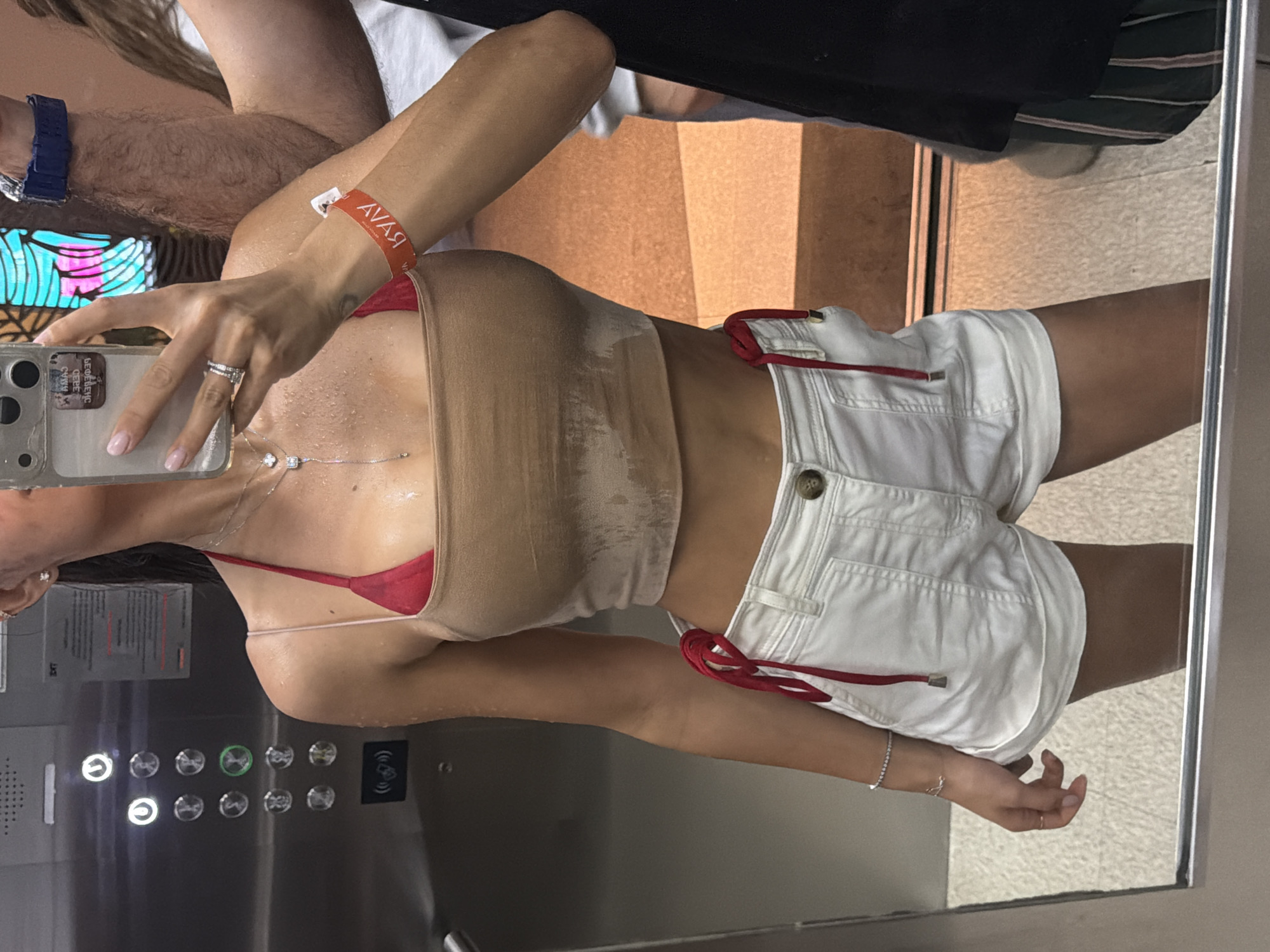A woman stands in an elevator, taking a mirror selfie. She wears a beige top over a red bikini top, white shorts with red ties, and has a red wristband on her left wrist. She holds her phone in her left hand, displaying rings on her fingers. A necklace is visible around her neck. The elevator panel with buttons is on the left. Reflections show another person behind her and a tiled floor outside the elevator. The wristband reads "AVAVA". The location is assumed to be Choeng Thale, Thailand.