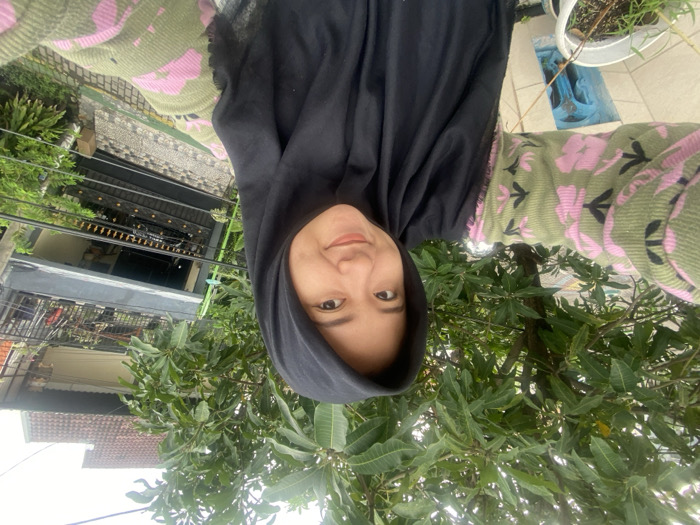 A person wearing a black hijab and a green and pink floral patterned top stands outdoors amidst green trees. The background includes a house with a brown roof, a balcony with a black railing, and other structures visible in the distance. The image is vertically inverted, placing the ground and background above the subject's head. A white potted plant sits on a tiled surface in the upper right corner.