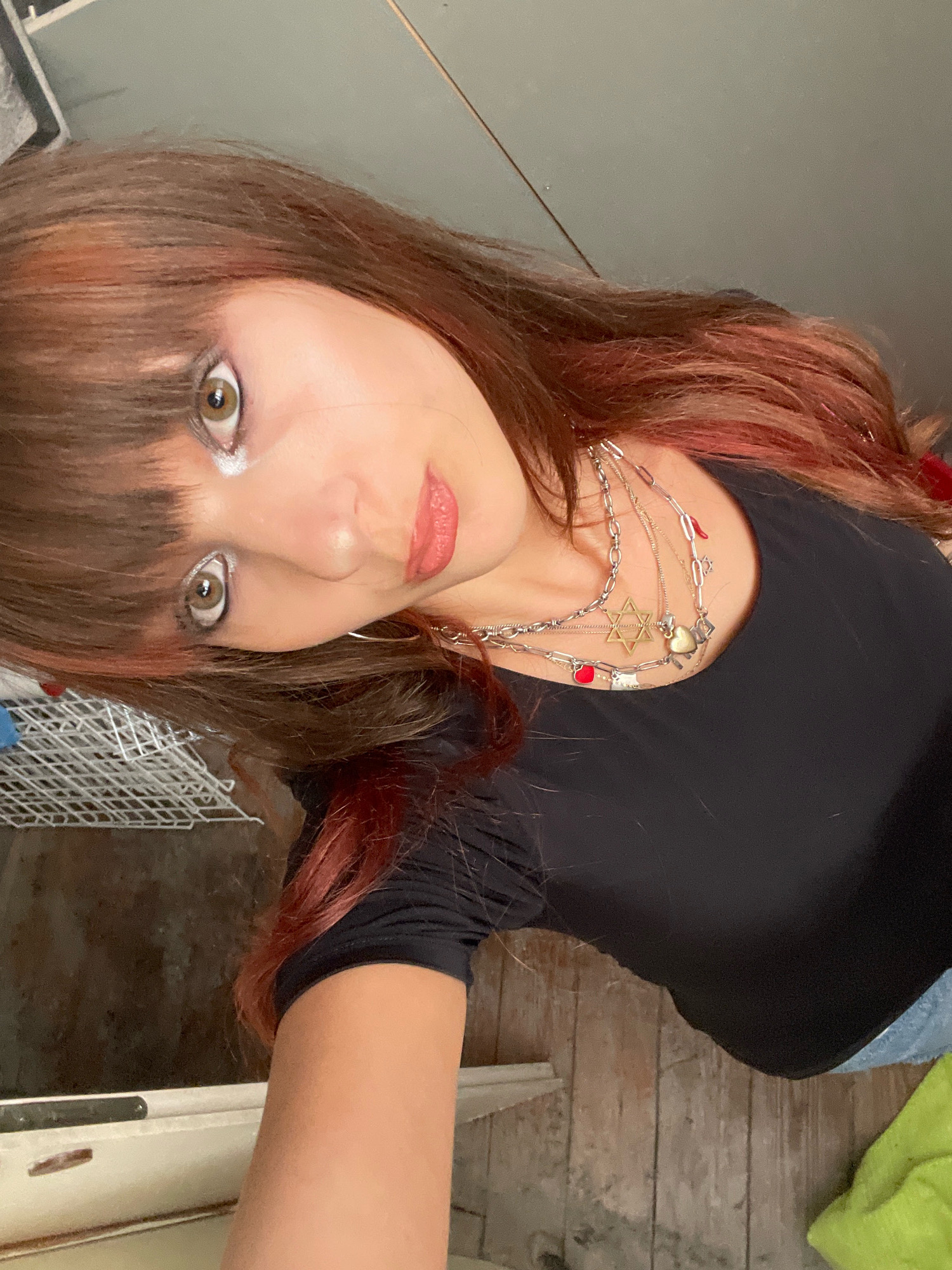 A young woman with reddish-brown hair and bangs is looking at the camera in a close-up, overhead shot. Her eyes are open and looking forward, with dramatic eye makeup. She is wearing a black t-shirt with multiple necklaces. One necklace features a Star of David pendant, while others have heart-shaped charms and other small pendants. The background appears to be an indoor environment with wooden floorboards and what looks like a shelf or counter. The image is not identifiable as being in Raanana, Israel.