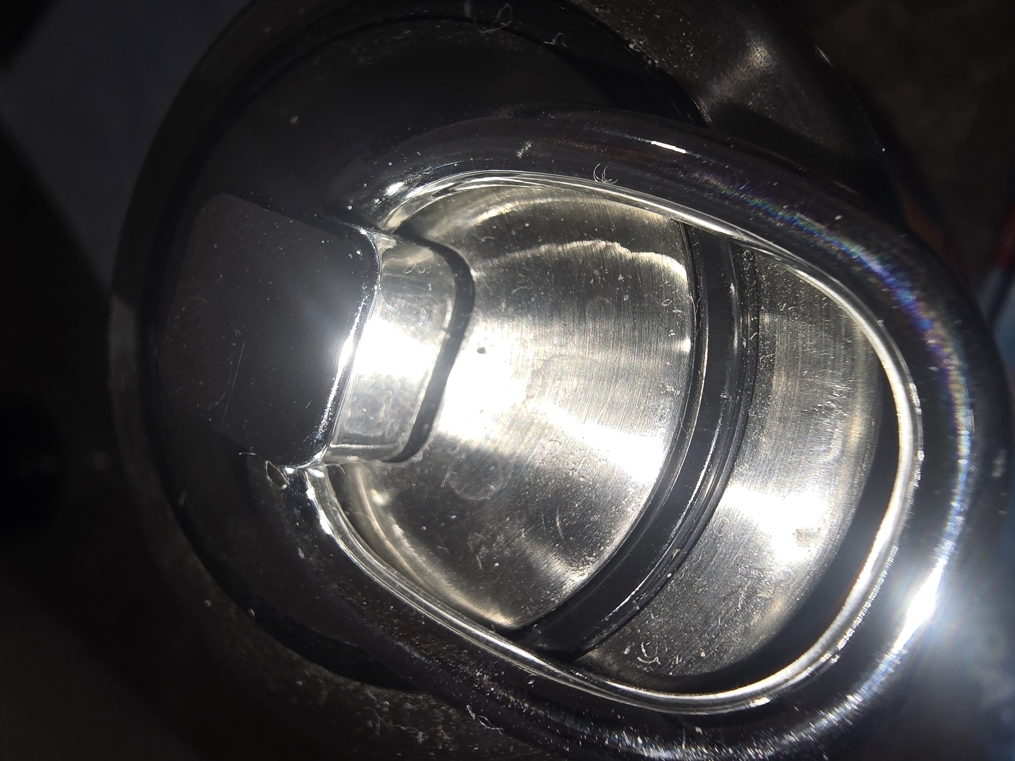 This image is a close-up, overhead shot of a metal handle mechanism, likely from a piece of luggage or a container. The handle is oval-shaped and made of polished, reflective metal, showing signs of wear and some light scuff marks. In the center of the oval is a raised, rectangular piece of black material, possibly plastic or rubber, which appears to be a locking or securing mechanism. The lighting is harsh and direct, causing significant glare and reflections on the metallic surfaces, with rainbow-like refractions visible on the lower right portion of the handle. The background is dark and out of focus, suggesting the object is indoors or in a dimly lit environment. There are no people or any identifiable features of the city of Ulan-Ude, Russia, present in the image.