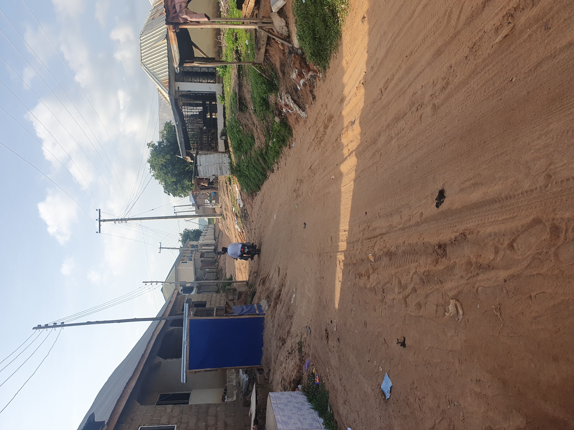 The image depicts a dirt road in Lokoja, Nigeria, with buildings lining both sides. The road is unpaved and shows tire tracks from vehicles. Power lines are visible overhead, supported by wooden poles. Several structures are present, including houses with corrugated metal roofs and some with cinder block construction. One building on the left has a blue tarpaulin covering a section. On the right, there are market stalls or open-fronted shops. A person is riding a motorcycle down the road away from the camera. There is debris scattered on the ground, including plastic bags and other refuse, indicating a lived-in environment. The sky is partly cloudy.