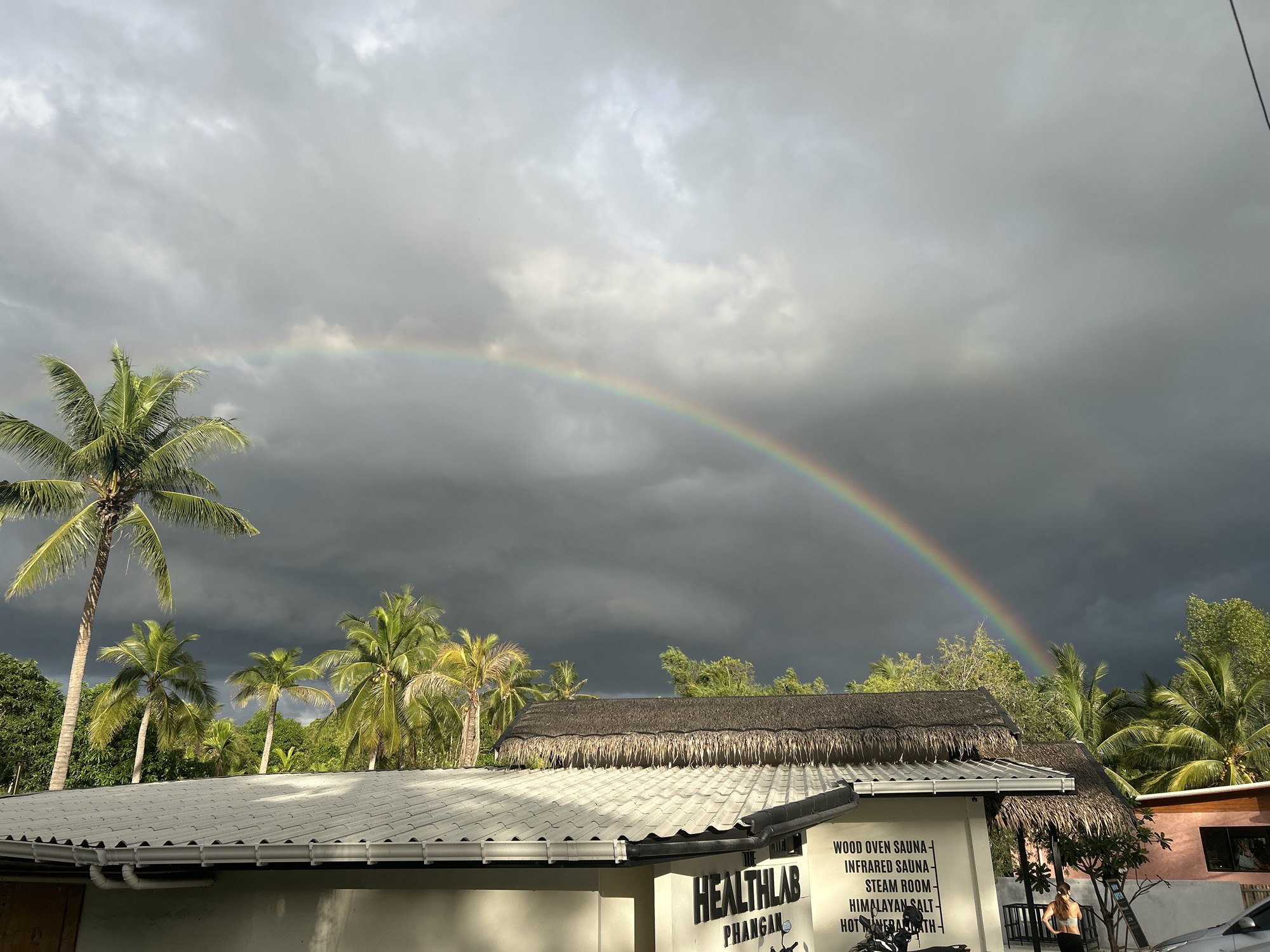 The image shows a view in Baan Hin Kong, Thailand, featuring a rainbow arching across a cloudy sky after a storm. In the foreground, there's a building with a tiled roof and white walls, bearing the name "HEALTHLAB PHANGAN" in bold black letters. Text on the side advertises various health treatments including "WOOD OVEN SAUNA", "INFRARED SAUNA", "STEAM ROOM", "HIMALAYAN SALT-", and "HOT HERBAL BATH-". A thatched roof structure is adjacent to the building. Palm trees are visible amidst lush green vegetation in the middle ground. A woman stands beside the building in a sports bra and leggings, looking towards the trees.