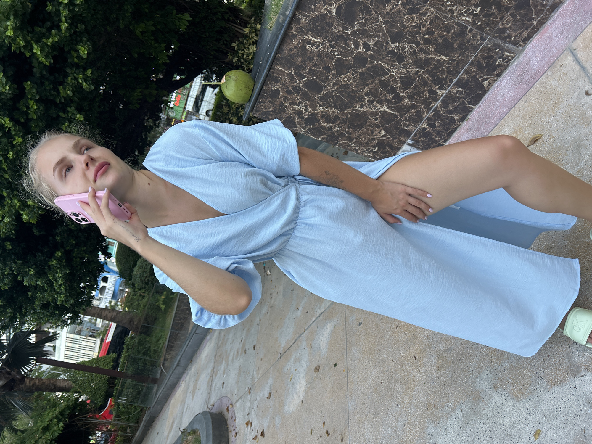 A young woman with blonde hair is sitting outside, talking on a pink smartphone. She is wearing a light blue, flowy dress that reaches her knees. Her right hand is holding the phone to her ear, and her left hand is resting on her thigh. The woman is looking upwards and to her right, with a relaxed expression.
The setting appears to be a park or outdoor seating area. Behind her, a tree with dark green leaves provides shade. To her right, there is a marbled surface and a light-colored, textured ground. A green coconut is visible in the background, hanging from a branch. In the further background, there are glimpses of buildings and vegetation, suggesting an urban or resort area. The lighting suggests it is daytime, with bright, natural light. The overall atmosphere is casual and relaxed.