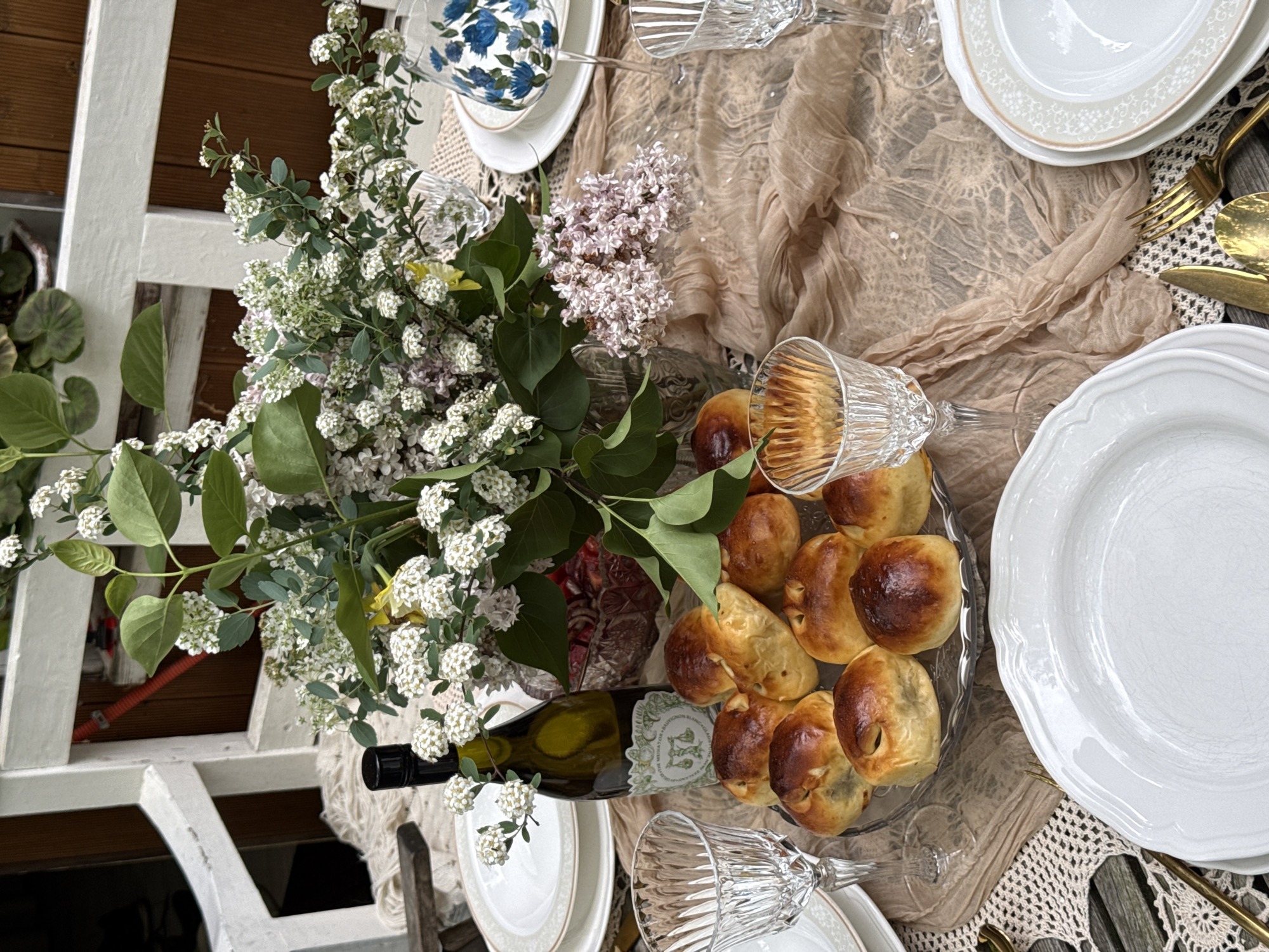 The image depicts a tablescape set for a meal. A white wine bottle is placed alongside a platter of pastries. A large bouquet of white and purple flowers occupies the center of the table. Wine glasses are laid on their sides near the pastries and plates. The plates are mostly white with some gold trim and are situated next to gold-colored silverware. The table is covered with a beige-colored, lacy cloth. The setting appears to be outdoors, near a white wooden structure. There is no indication that this is in Sapronovo, Russia.